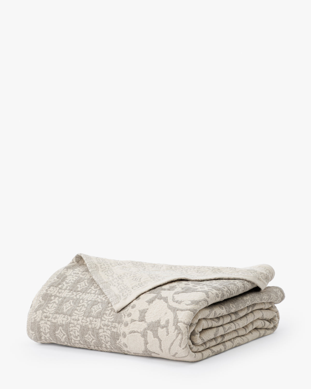 The Floral Matelasse Coverlet, featuring folded beige and light gray fabric with subtle floral and geometric patterns, is displayed on a plain white background.