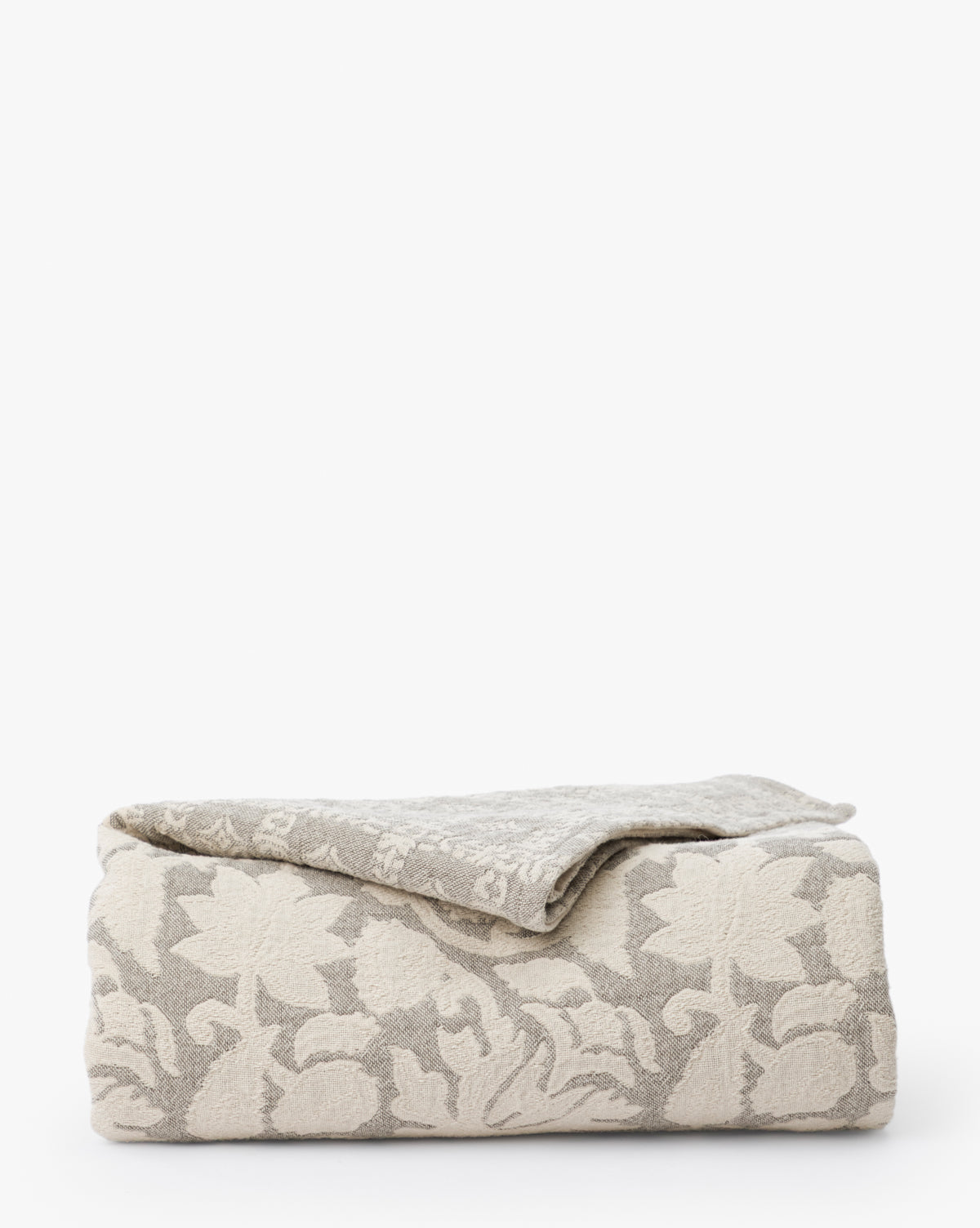 The Floral Matelasse Coverlet, in folded beige and light gray cotton with a raised floral and leaf pattern, evokes elegant matelasse designs and is shown against a plain white background.