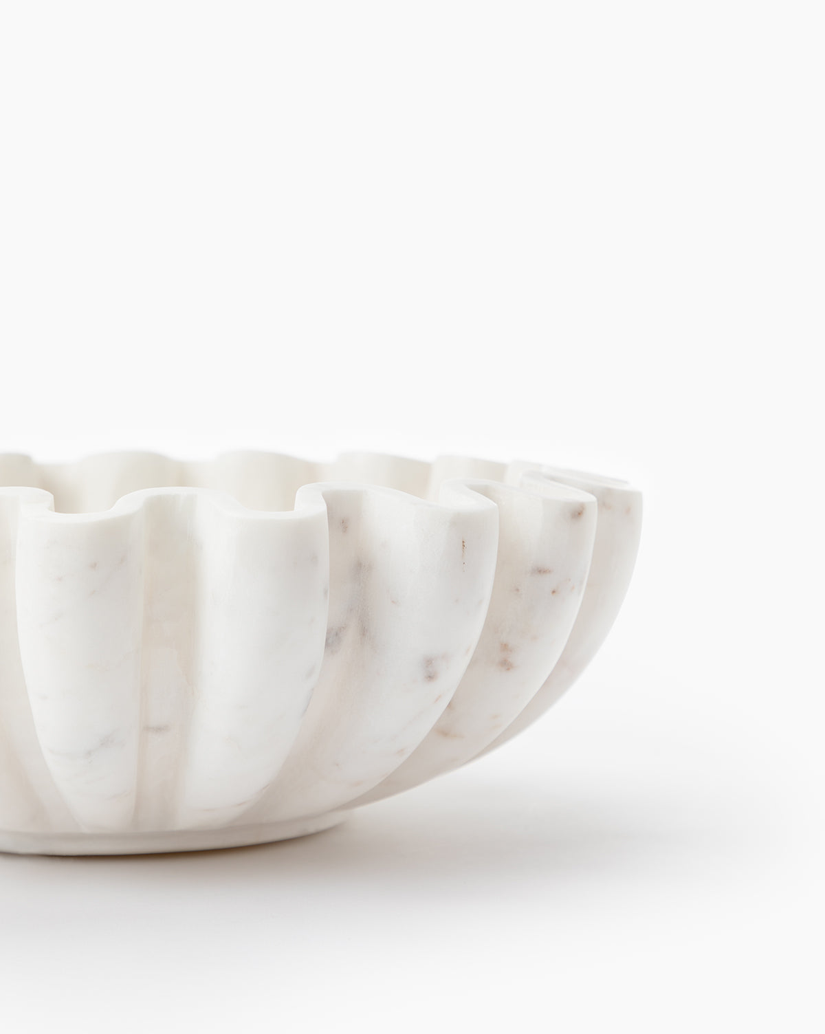 A close-up of the Fluted Marble Bowl, featuring its scalloped edge and elegant carving, is shown against a plain white background, with only half of the bowl visible in the image.