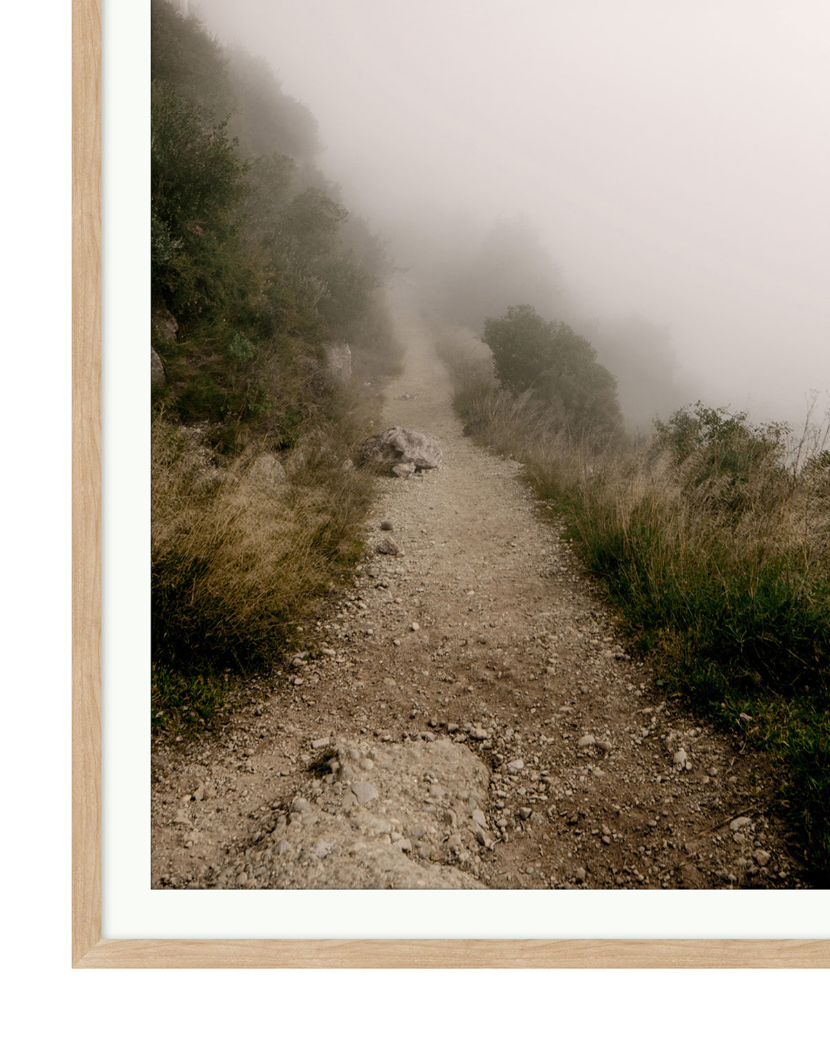 A narrow dirt path winds through grassy hills into thick fog, surrounded by bushes and rocks, with a light wooden frame bordering the Foggy Path giclee print.