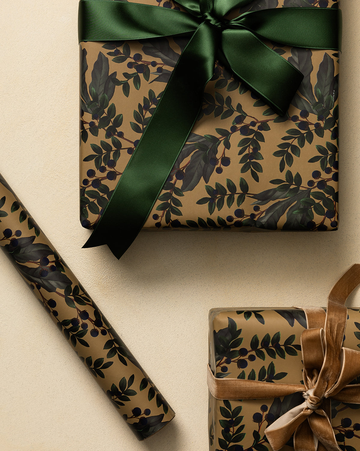 Two gifts wrapped in Foragers Gift Wrap, featuring brown floral patterns with green leaves and dark berries: one tied with a green satin ribbon, the other with brown velvet. A matching roll of Foragers Gift Wrap rests nearby on a beige surface, by McGee & Co.