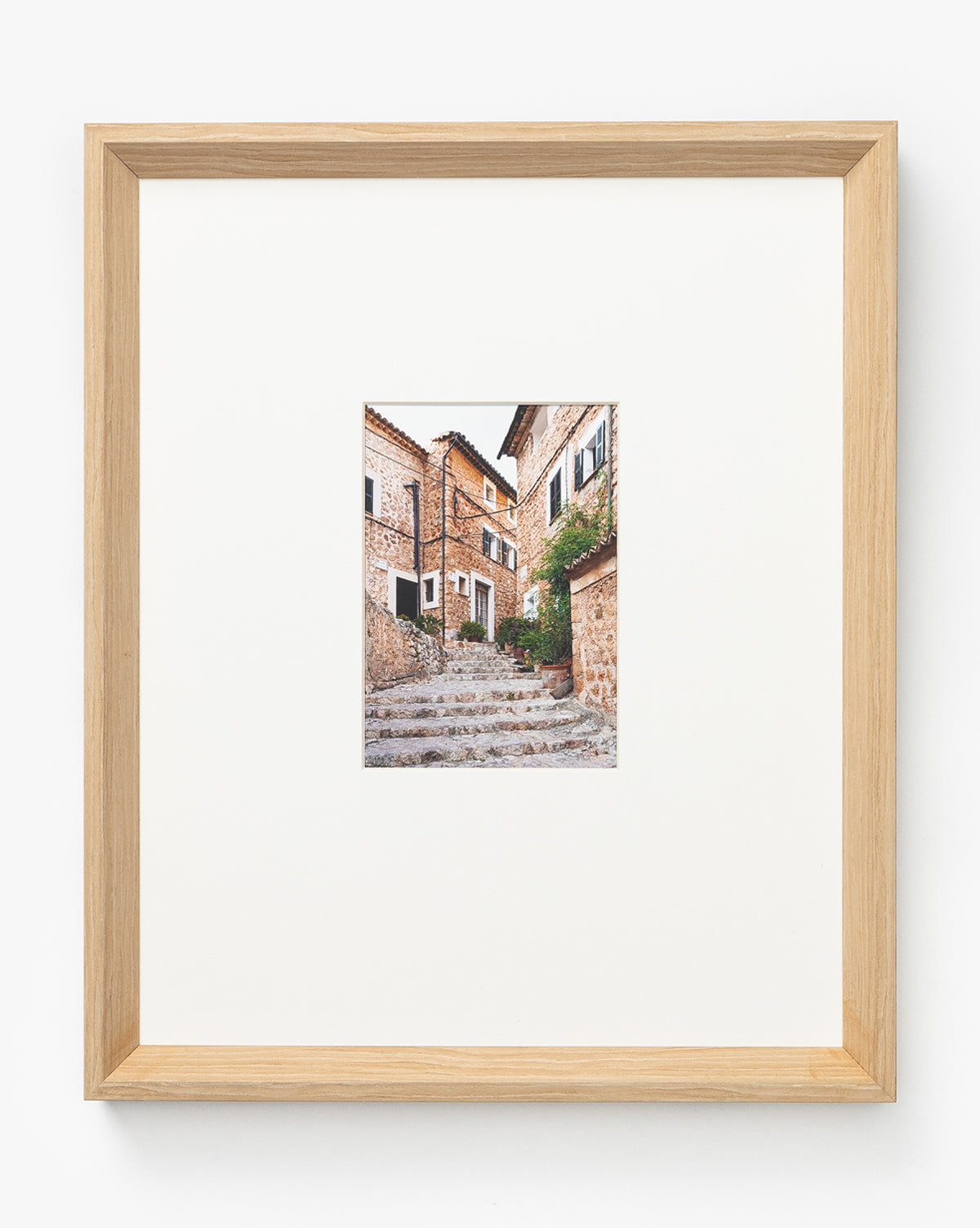 Featuring Fornalutx, this stunning giclee print captures a stone alleyway with stairs, rustic brick buildings, and potted plants—centered on a white background in a light natural woodgrain frame, perfect for any room.