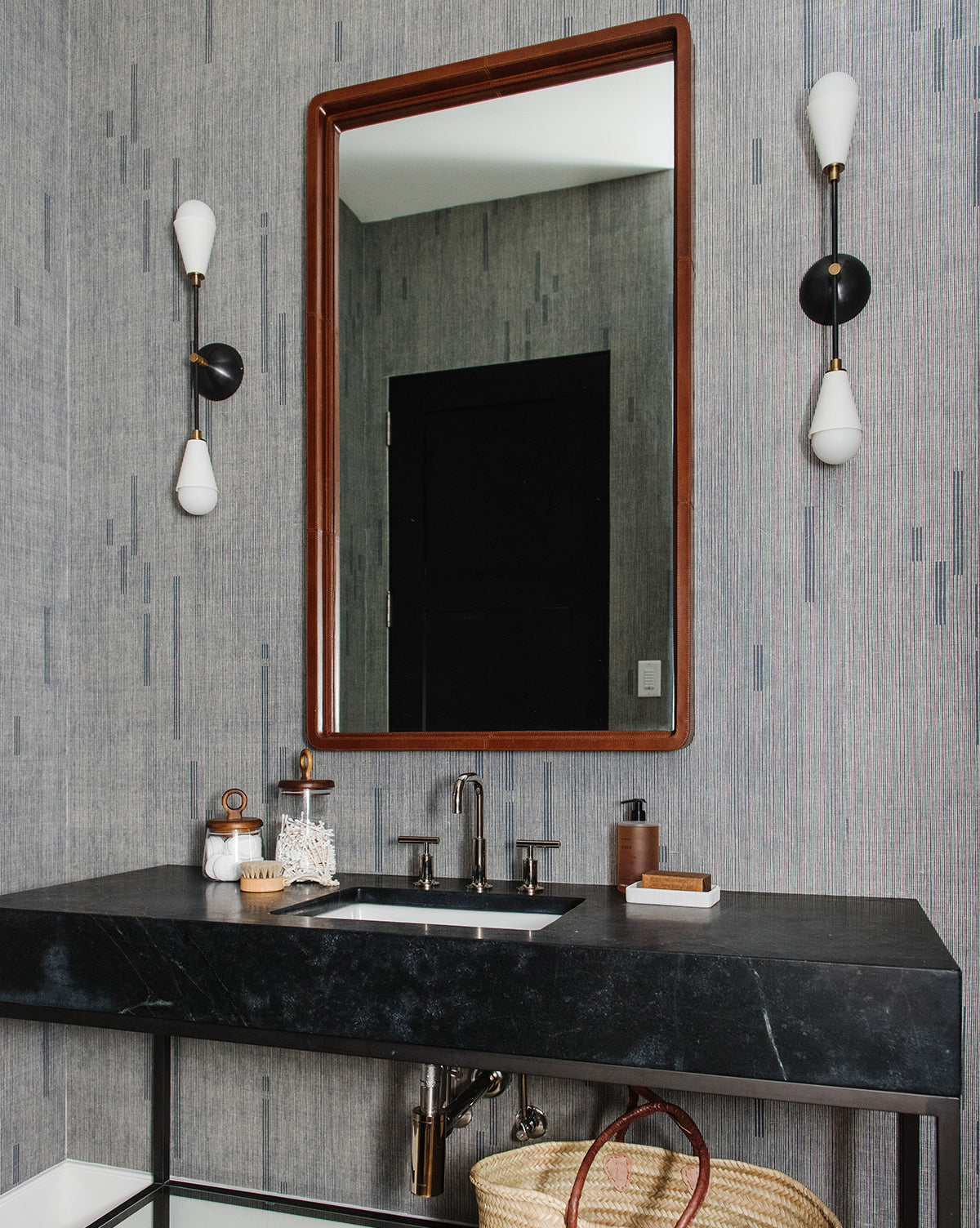 A modern bathroom features a black countertop, rectangular sink, and chrome faucet. Above hangs the Franklin Leather Mirror, flanked by black and white sconces. A woven basket sits under the sink, by McGee & Co.
