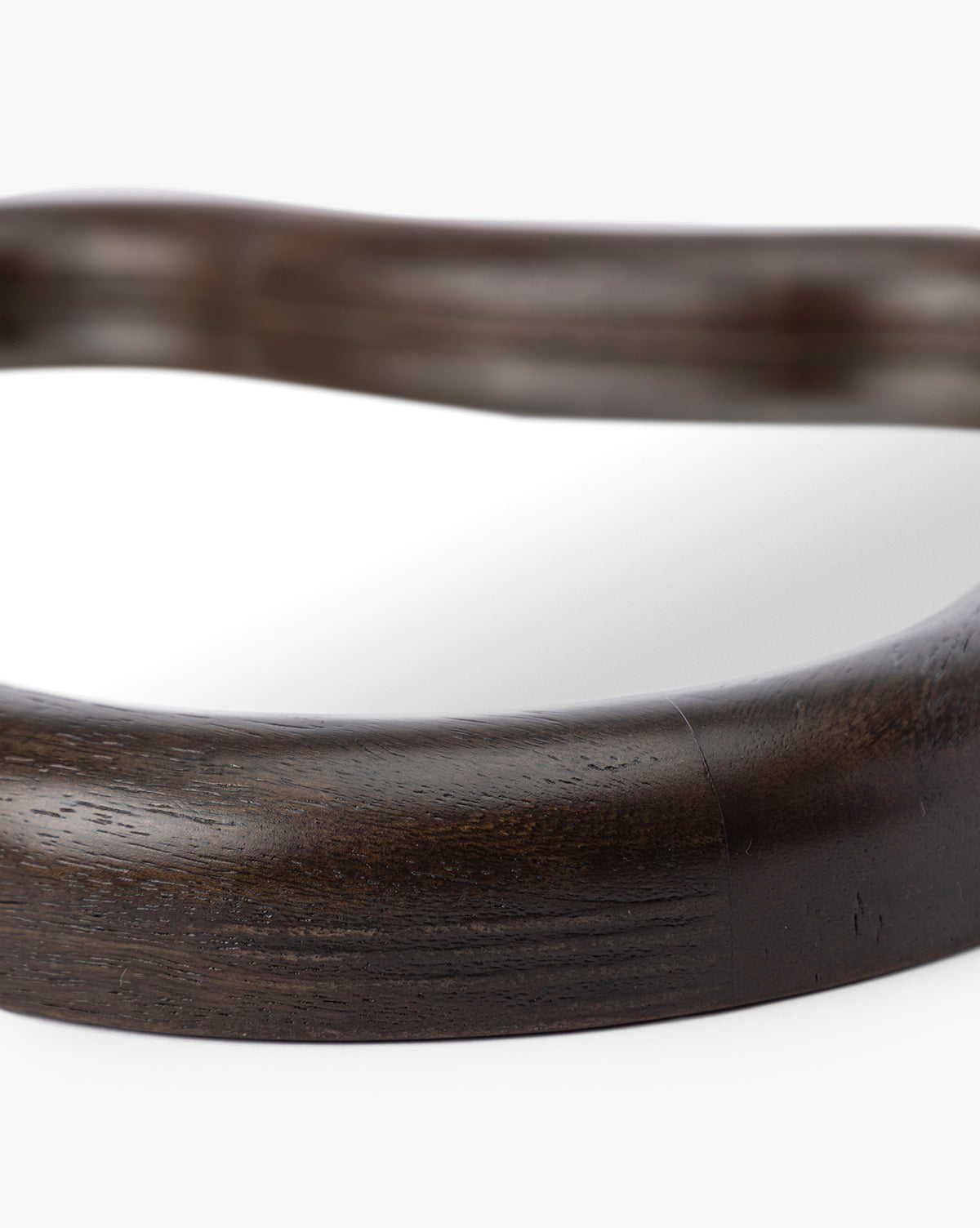 Close-up of the Freya Shelf Mirror’s dark, smooth, curved wooden frame with visible wood grain against a white background—an elegant entryway accent with part of the reflective surface shown by McGee & Co.