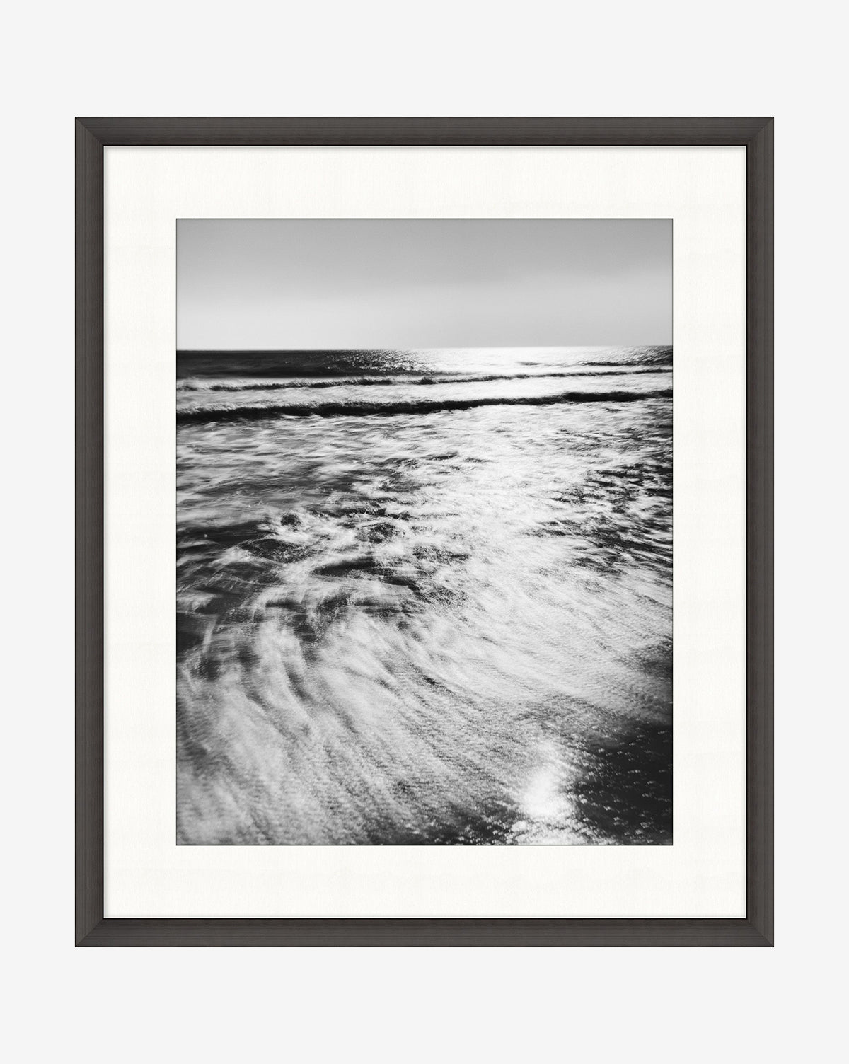 Frozen Waves is a black and white photograph of ocean waves rolling onto shore, sunlight reflecting on the water. This elegant coastline wall art comes framed in satin charcoal with a crisp white mat.