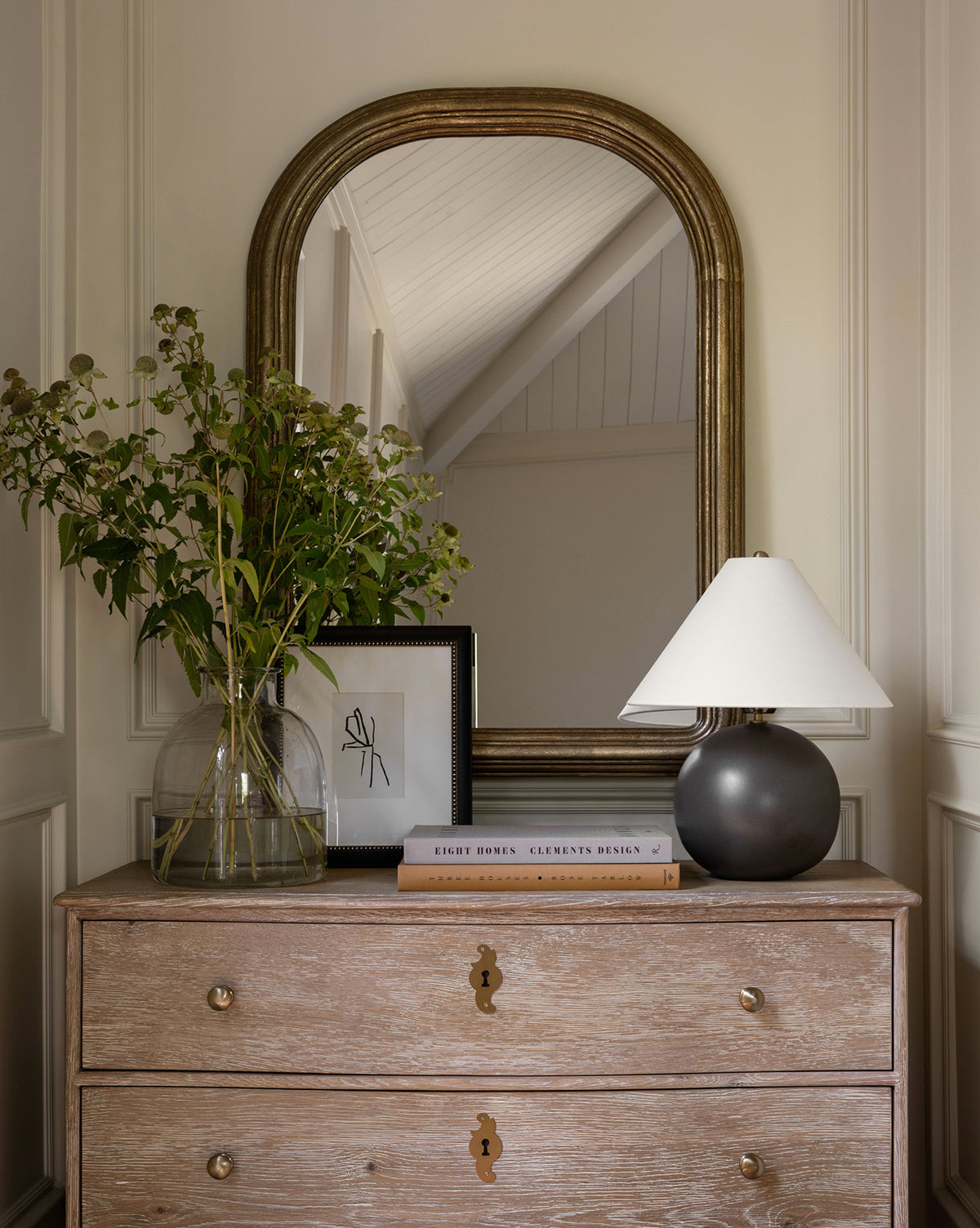 A wooden dresser with two drawers holds a glass vase of green foliage, a small framed sketch, stacked books, and the Tess Ceramic Table Lamp with a white shade; an arched mirror above reflects the ceiling, by McGee & Co.