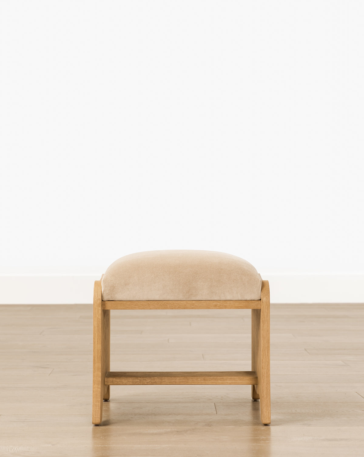 The Makerspalm Gable Ottoman, a small cushioned seat in fawn velvet with a drift oak frame, rests on a light brown wooden floor against a plain white wall—McGee & Co.