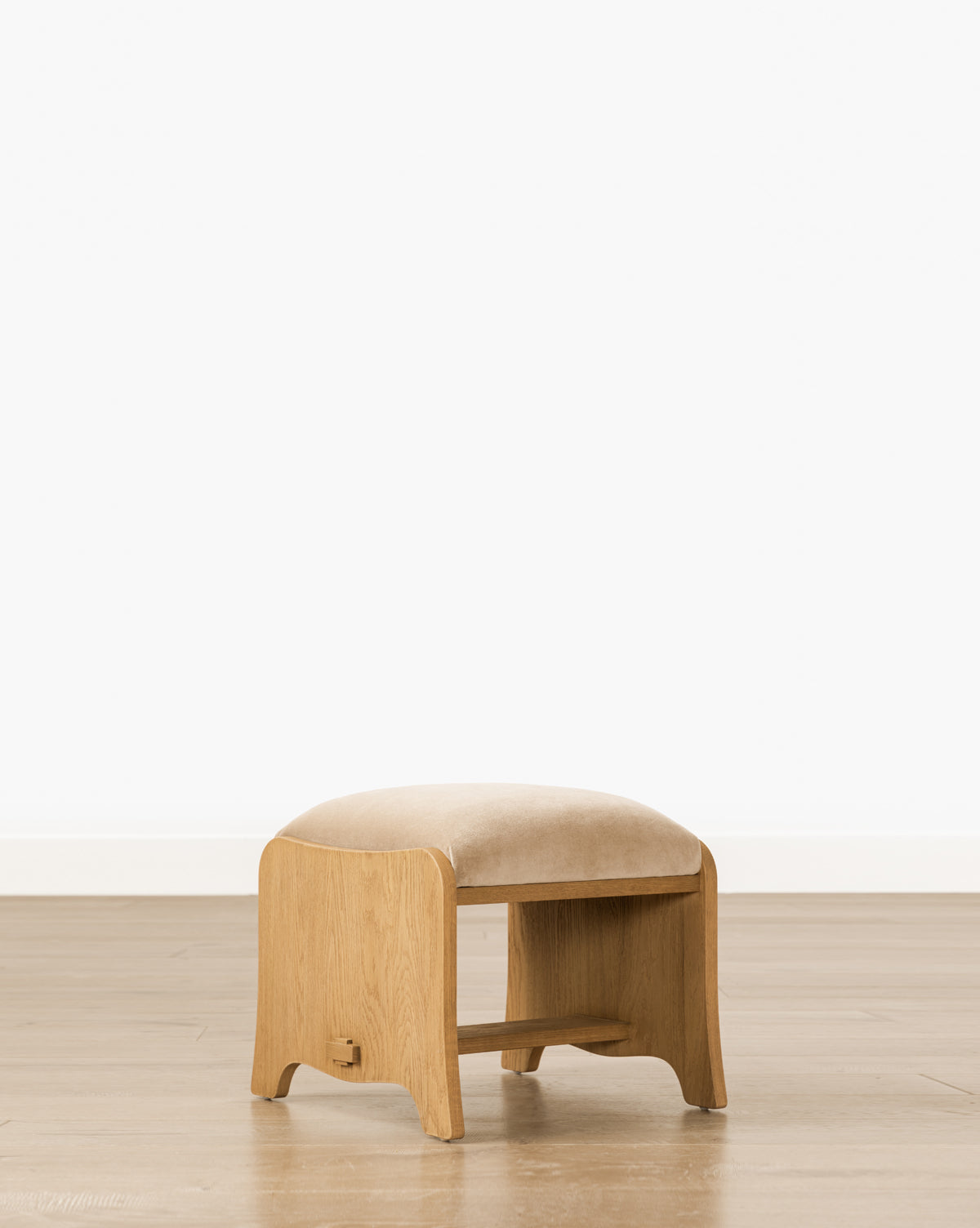 The Makerspalm Gable Ottoman features a Drift Oak frame, rounded edges, a low shelf, and a beige Fawn Velvet seat cushion. Styled simply, it stands on a wooden floor against a white wall. McGee & Co.