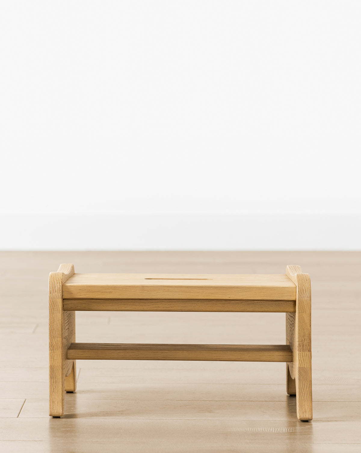 The Gable Step Stool by YANG CHENG WOODEN INDUSTRIES INTERNATIONAL (VIETNAM) CO LTD, made from drift oak with mortise and tenon joinery, features a rectangular top with rounded edges and is set on a wooden floor by a white wall. McGee & Co.