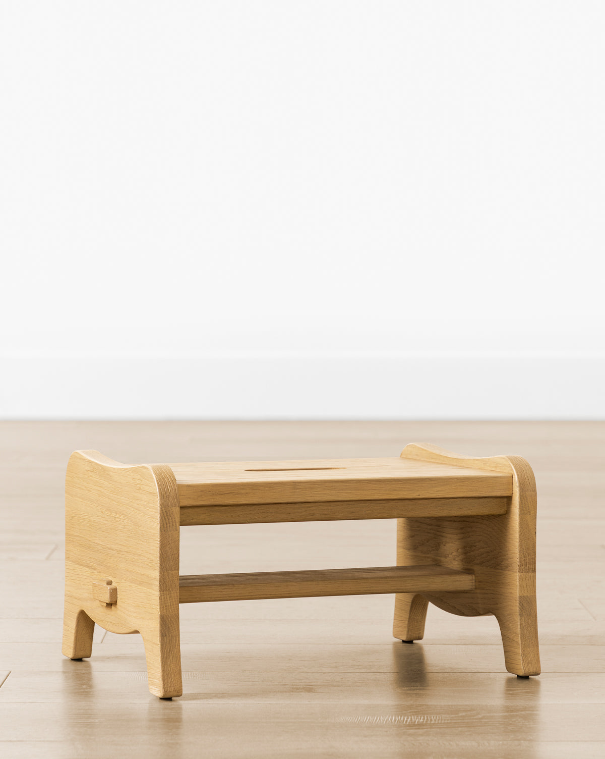The Gable Step Stool by YANG CHENG WOODEN INDUSTRIES INTERNATIONAL (VIETNAM) CO LTD, crafted in drift oak with curved edges, a central cut-out handle, and mortise and tenon joinery, stands on a light wooden floor against a white wall. McGee & Co.