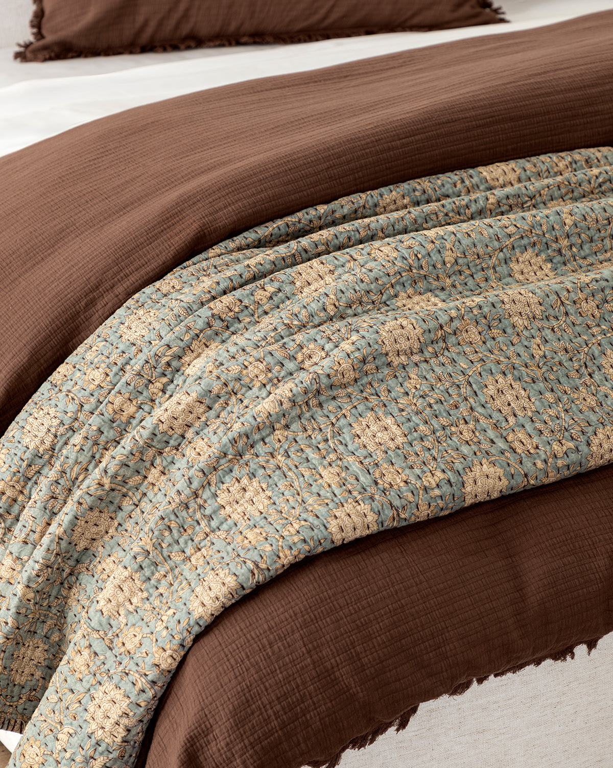 A close-up of a neatly made bed displays the Gabriella Block Print Quilt with blue and beige patterns, partially covered by a folded brown comforter. A matching brown pillow appears in the background.