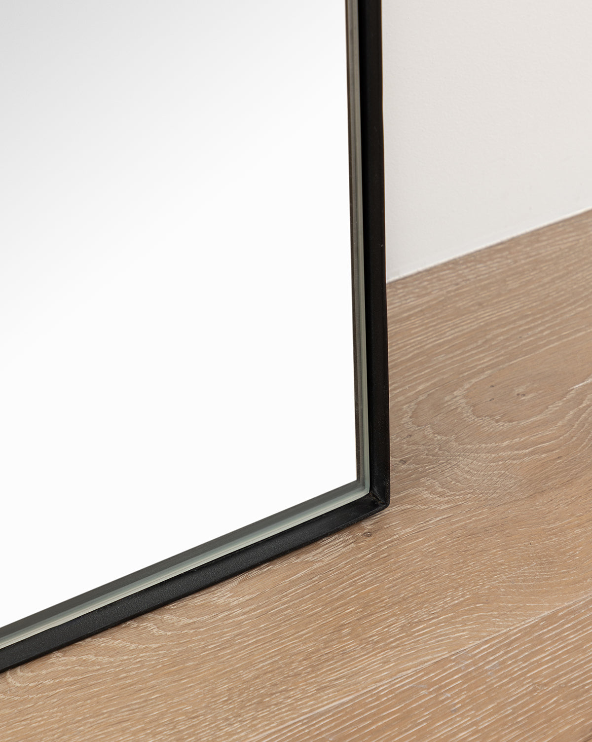 A close-up of the Gala Floor Mirror’s thin black frame resting on light wood beside a white wall—an ideal stylish addition to your bedroom mirror setup, by McGee & Co.