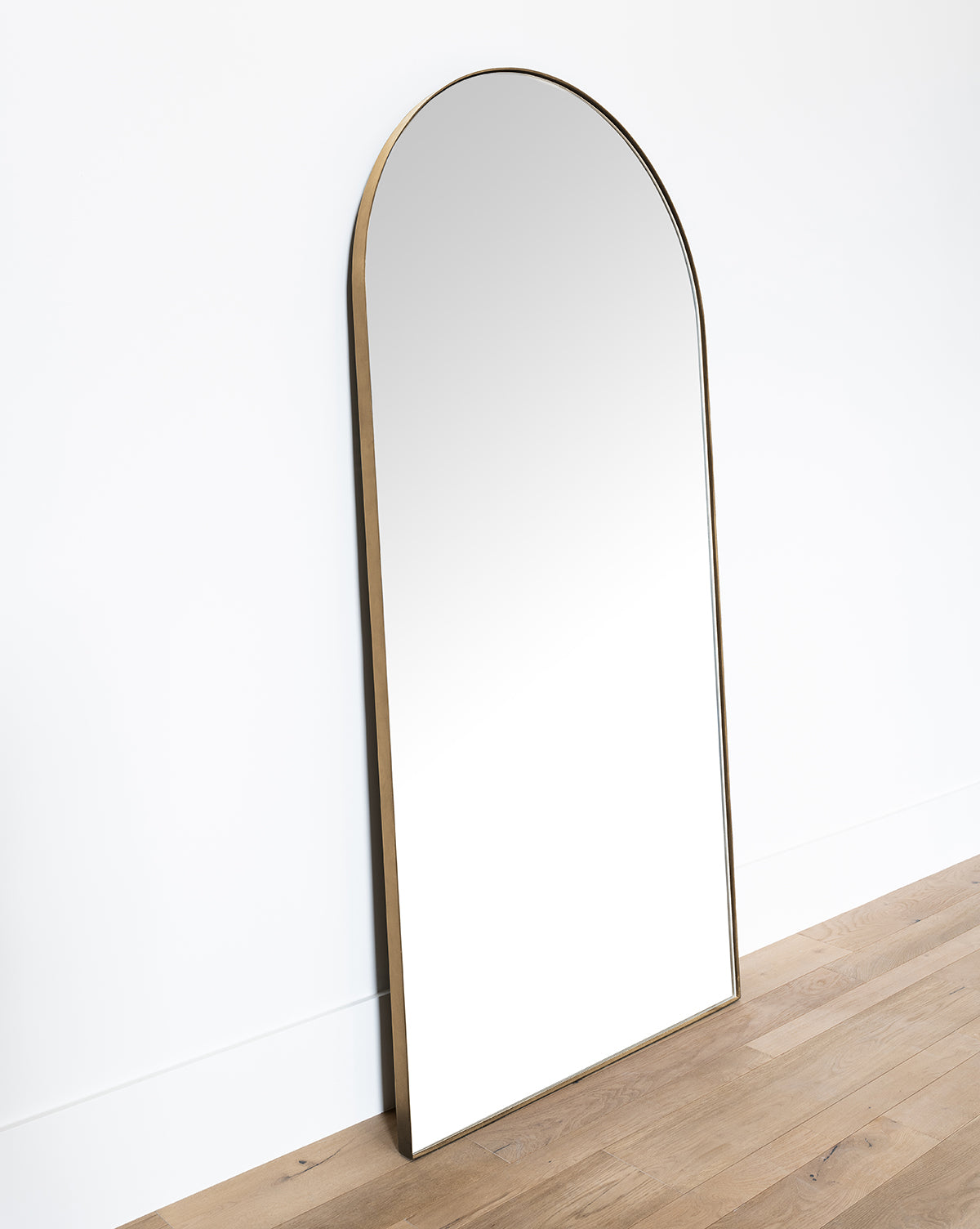 The Gala Floor Mirror features a large reflective surface and a slim gold frame, leaning against a white wall on light wood flooring. Its modern, minimalist design enhances any room with elegance and simplicity, by McGee & Co.