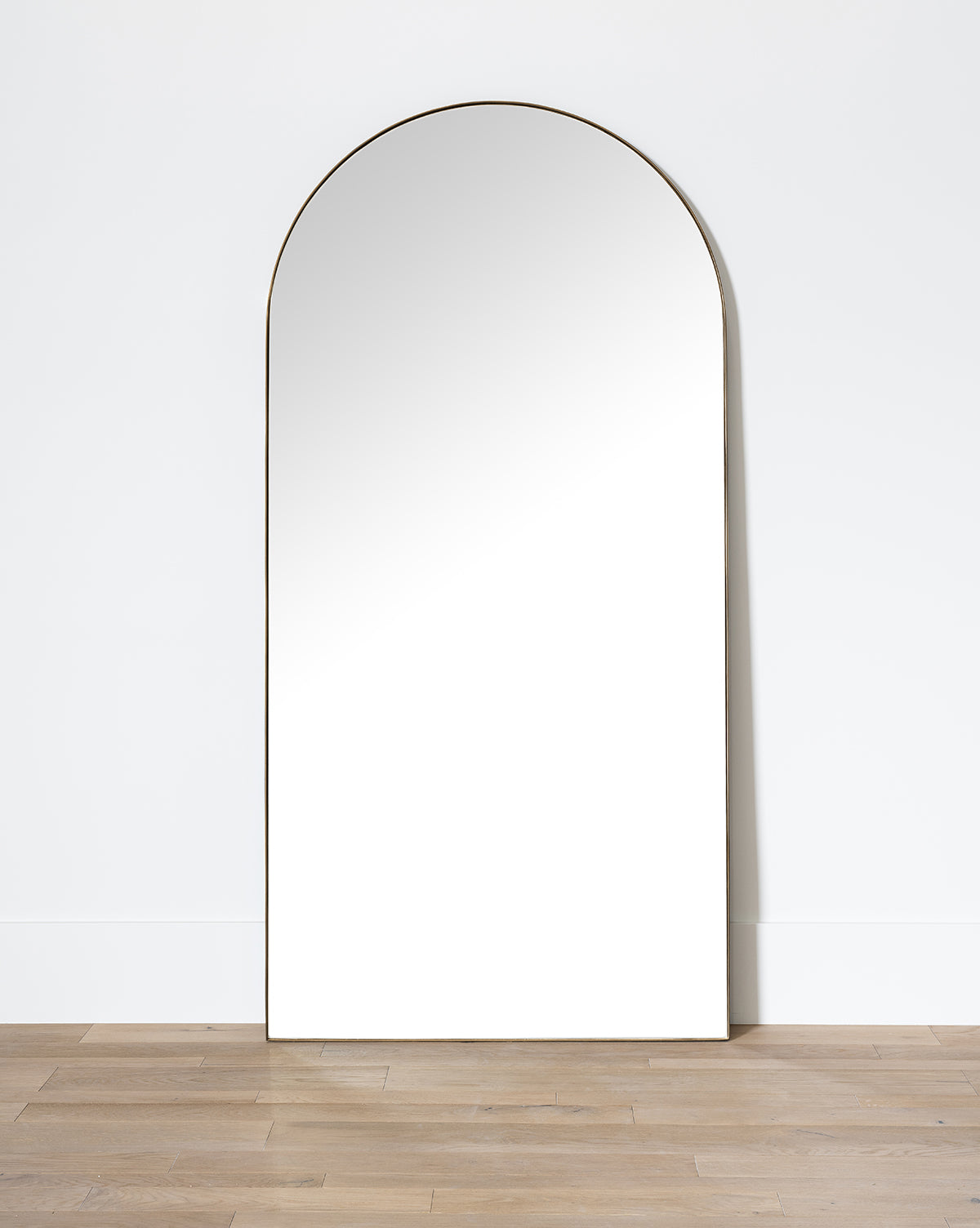 The Gala Floor Mirror, featuring a slim frame, is shown leaning against a white wall on a light wood floor, its reflection capturing the simplicity of the wall across from it, by McGee & Co.
