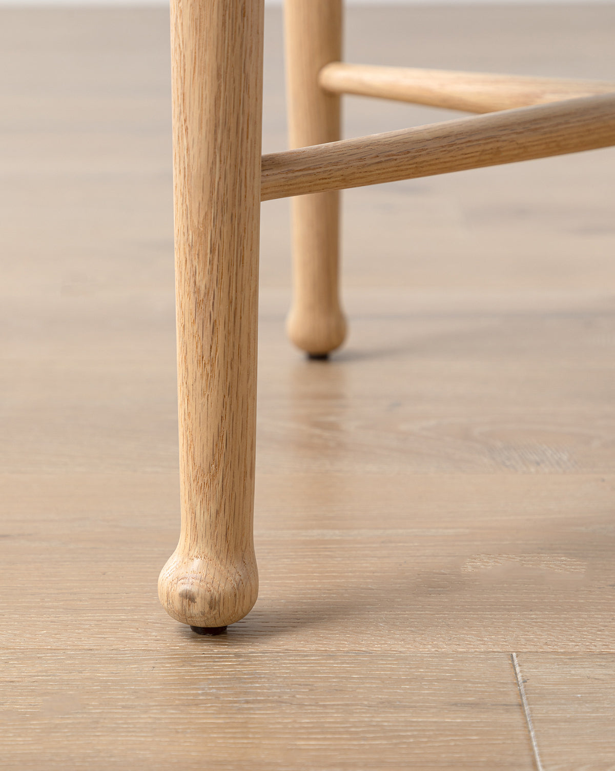 Close-up of the Garance Dining Chairs white oak leg with a rounded foot on a light wood floor. Another leg appears in the background, showcasing the chair’s smooth texture and natural grain, by McGee & Co.