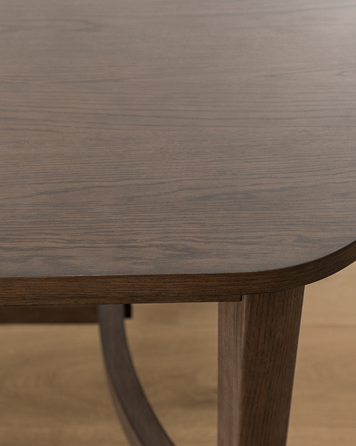Close-up of the Gardiner Dining Table reveals a rounded corner, visible wood grain, and a single leg. The table features a smooth Walnut Oak finish, contrasting with the lighter-colored wooden floor beneath, by McGee & Co.