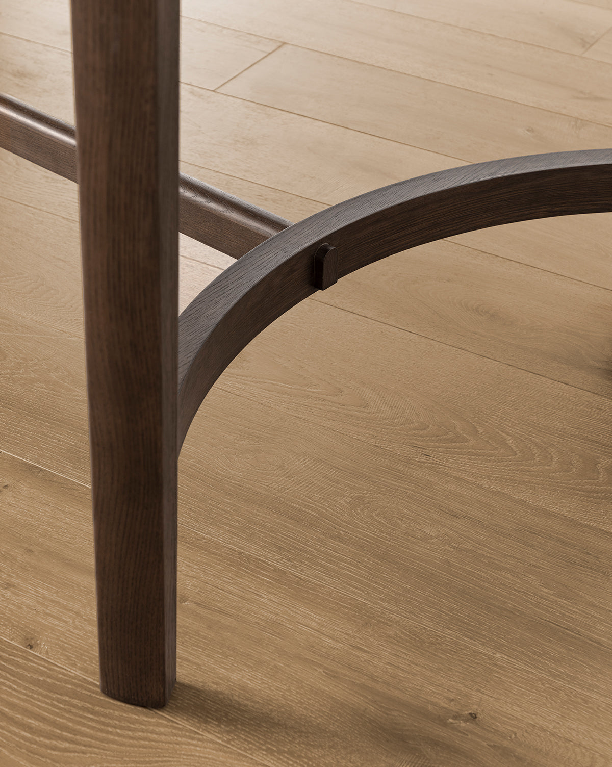 Close-up of the Gardiner Dining Table legs and curved support beam on a light wood floor, highlighting the Walnut Oak grain and texture featured in both the table and flooring, by McGee & Co.
