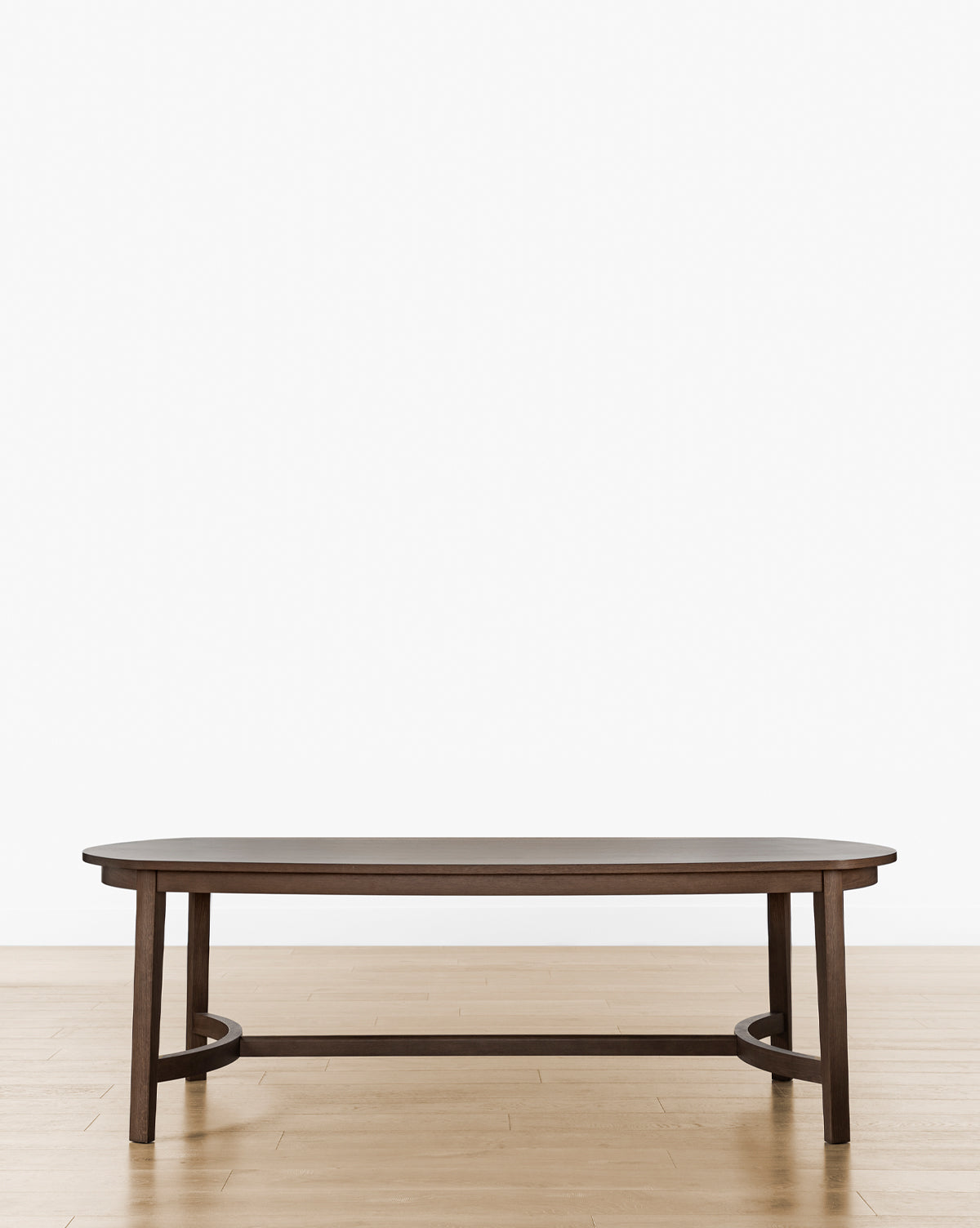 The Gardiner Dining Table, featuring a dark walnut oak oval design with four legs and a low crossbar base, stands elegantly on a light wood floor against a plain white background, by McGee & Co.