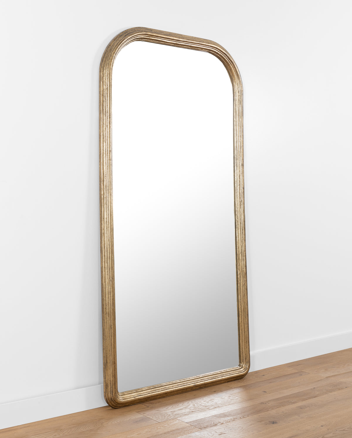 The Giada Floor Mirror, featuring a large arched design and elegant gold frame, leans against a white wall on a wooden floor, by McGee & Co.