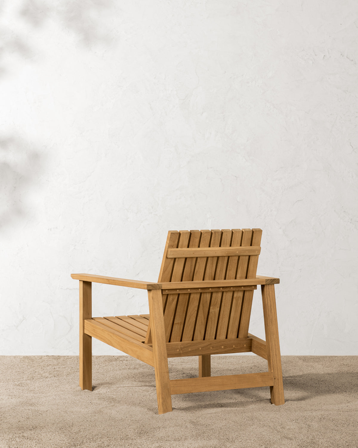 The Gilda Outdoor Lounge Chair, crafted from teak wood with vertical slats, sits on a gravel surface against a textured white wall by McGee & Co.