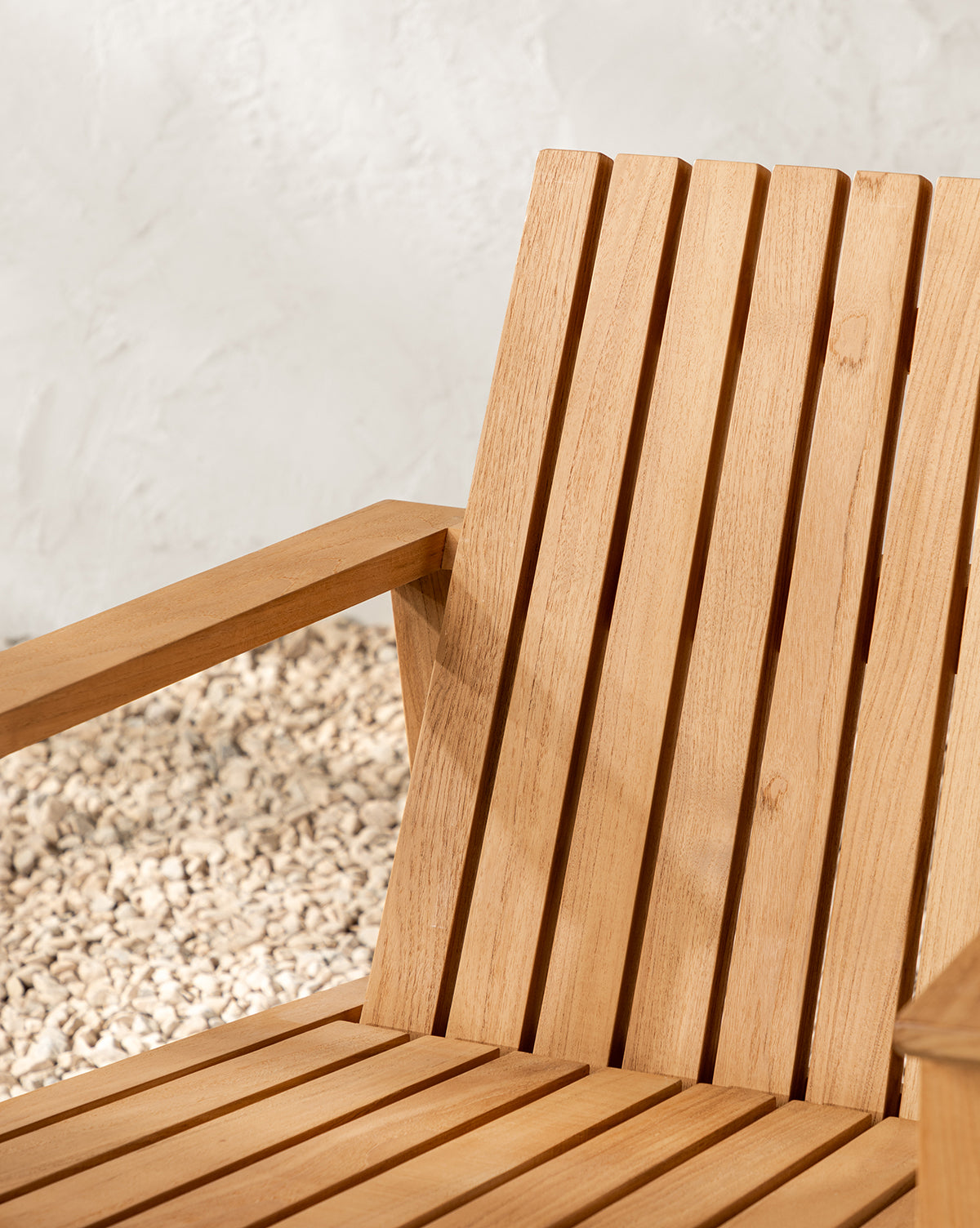Close-up of the Gilda Outdoor Lounge Chair made from teak wood, featuring vertical slats on the backrest and seat, angled armrests, and set against a backdrop of light pebbles and a textured white wall, by McGee & Co.
