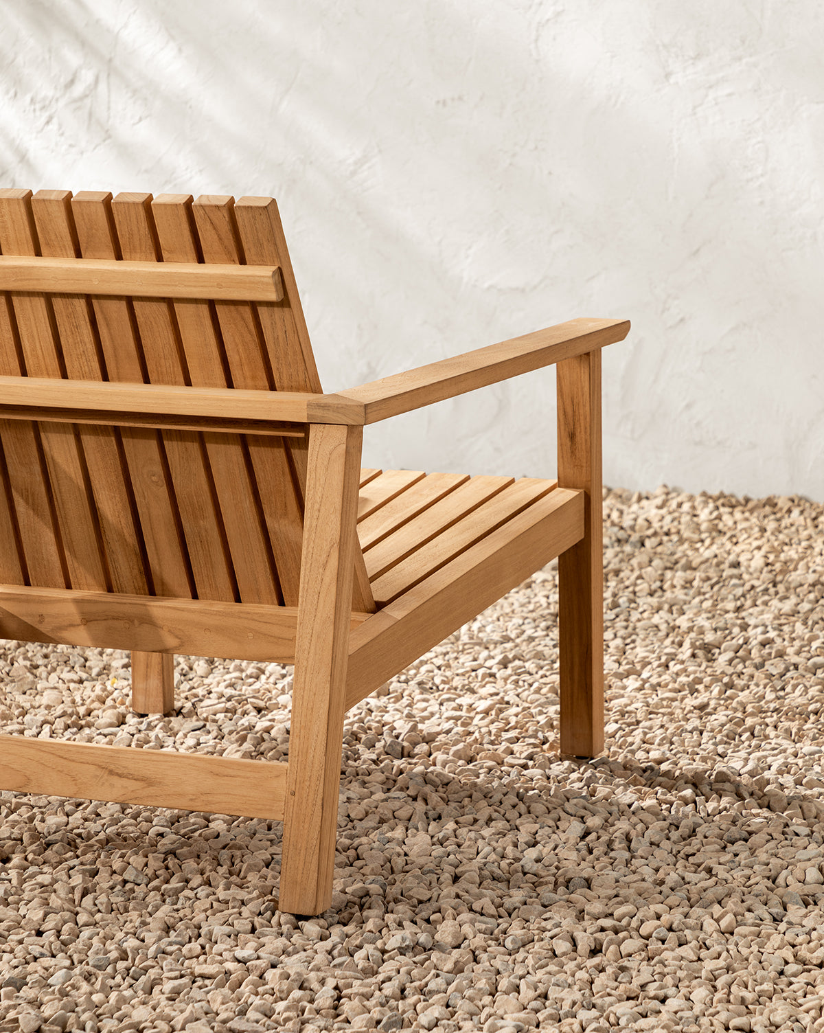 The Gilda Outdoor Lounge Chair, crafted from teak wood with a slatted back and seat, rests on small light-colored stones in front of a textured white wall, by McGee & Co.