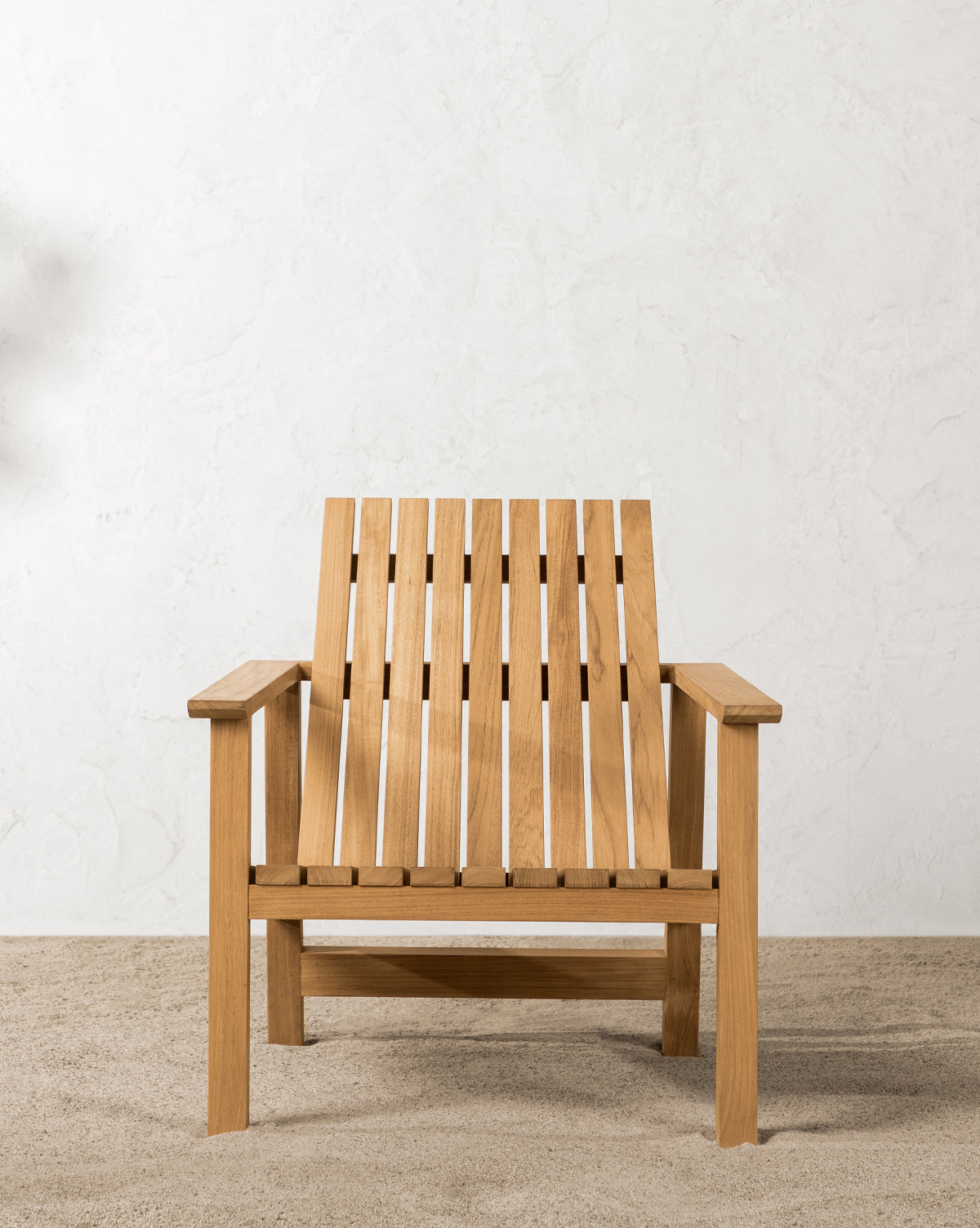 The Gilda Outdoor Lounge Chair, made of teak wood with a slatted seat and backrest, sits on gravel against a textured white wall in soft natural light, by McGee & Co.
