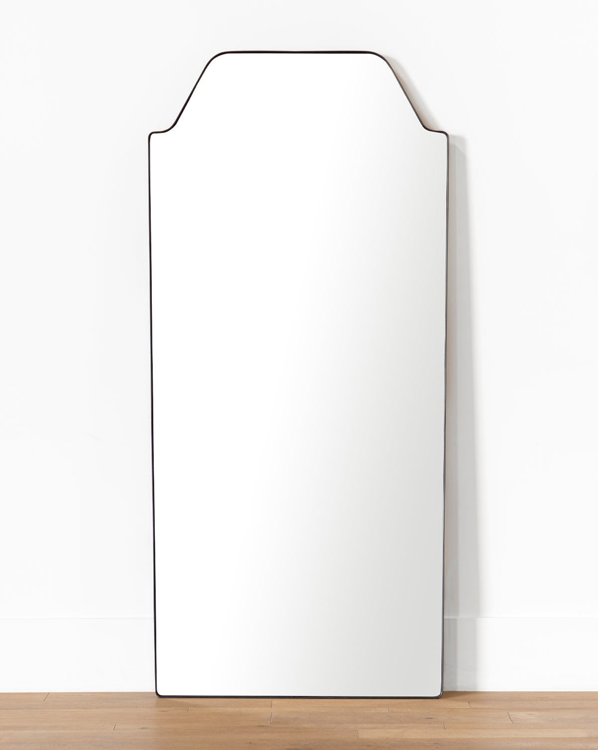 The Giles Floor Mirror, featuring a tall rectangular shape, unique notched top edge, and sleek matte black frame, stands on a wooden floor against a plain white wall by McGee & Co.