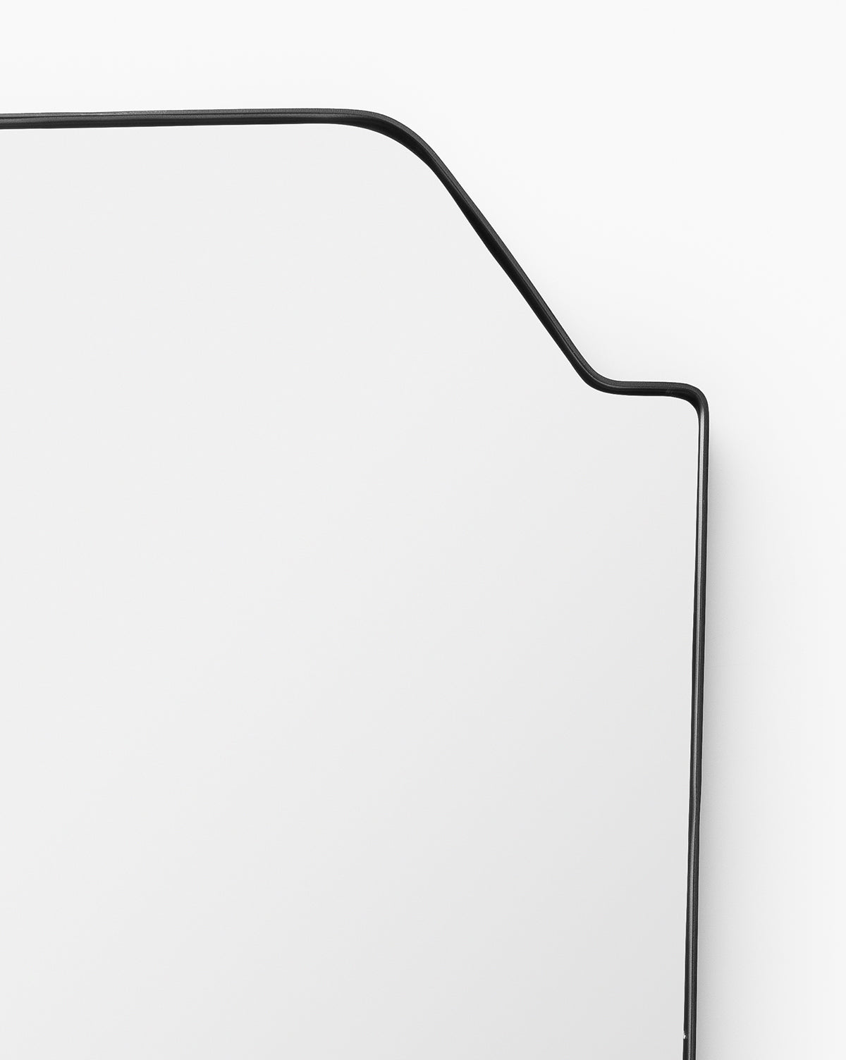 A close-up of the Giles Wall Mirror highlights its thin, matte black asymmetric frame bending at an angle, creating a striking modern geometric shape against a plain white background, by McGee & Co.