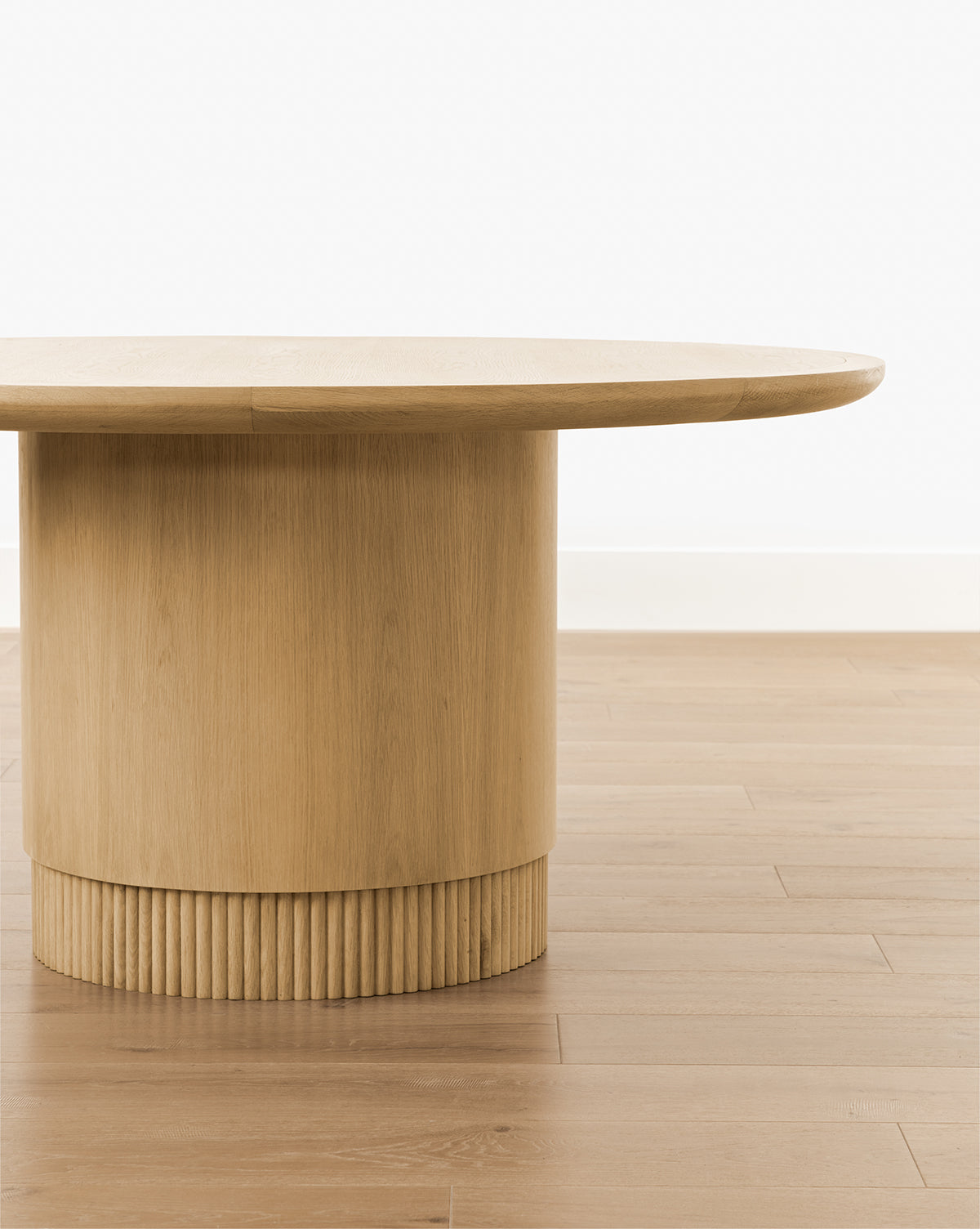 The Gilroy Round Oak Dining Table with a Heritage Oak Finish features a modern round top and a reeded cylindrical base with elegant vertical grooves, shown here on a light wood floor against a plain white wall, by McGee & Co.