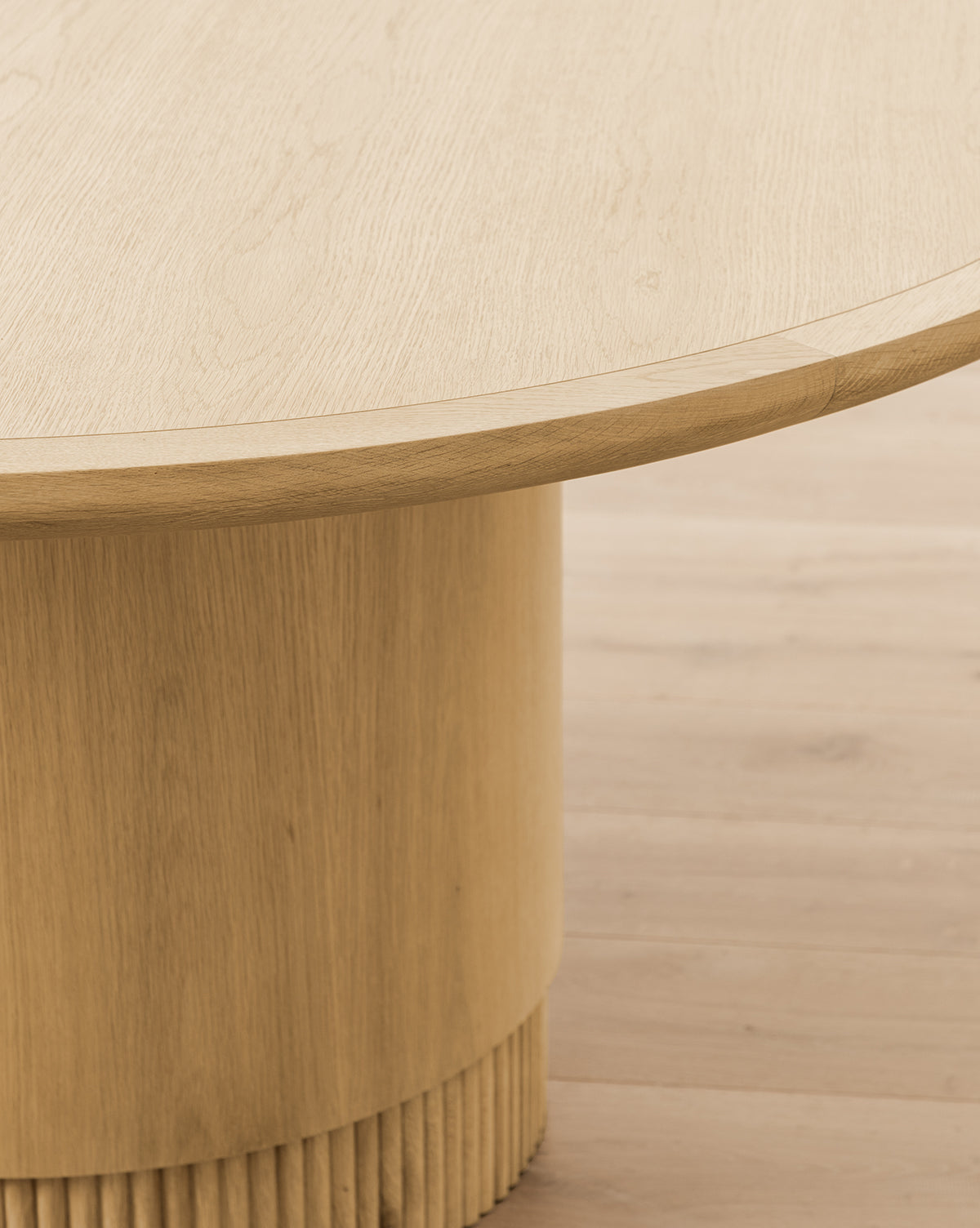 A close-up view of the Gilroy Round Oak Dining Table with a wide reeded base, set on a matching light wood floor. The smooth table surface and base display natural wood grain in a stunning Heritage Oak Finish, by McGee & Co.
