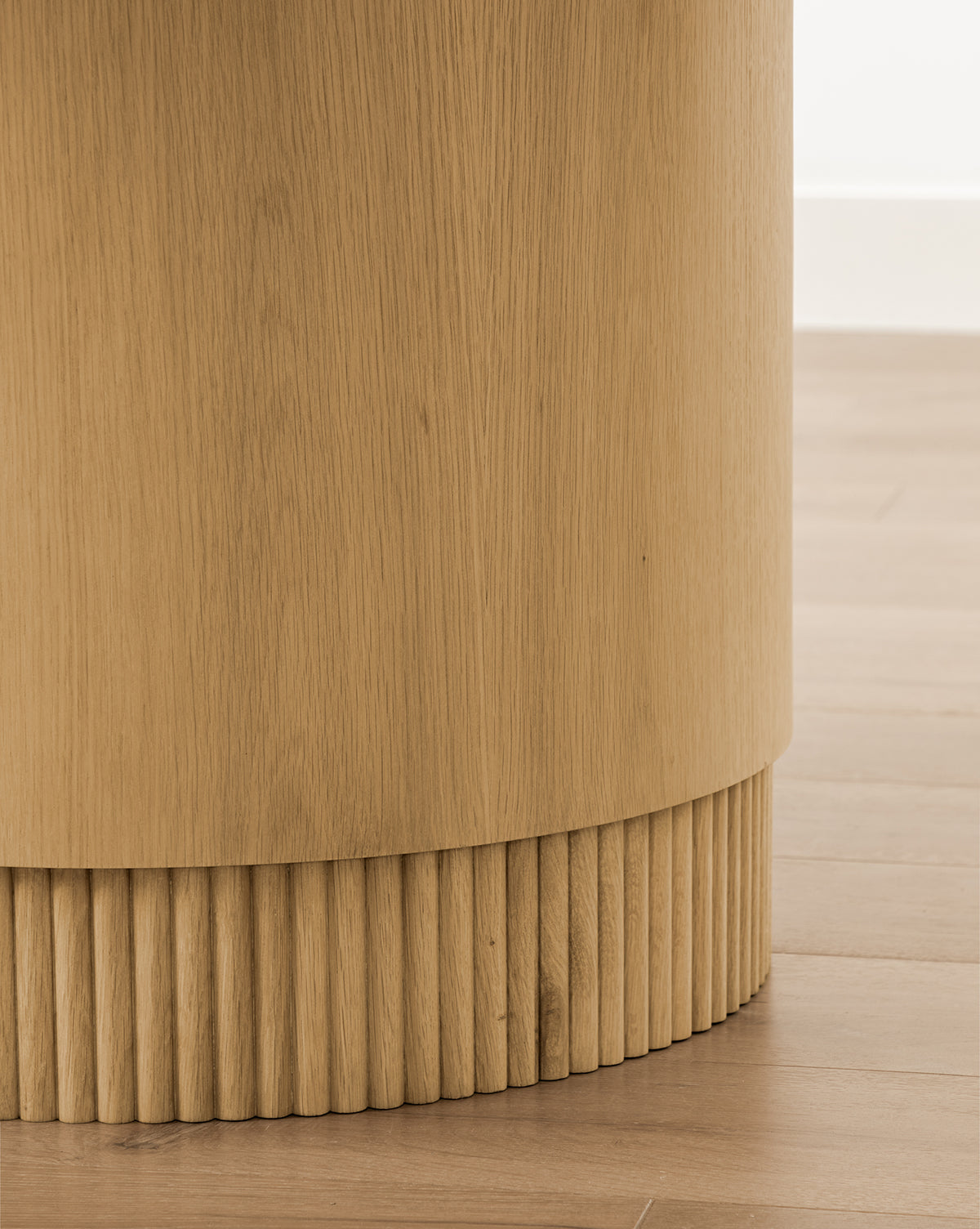 Close-up of the Gilroy Round Oak Dining Tables smooth cylindrical base with reeded detailing, shown on a light wood floor against a white wall, by McGee & Co.