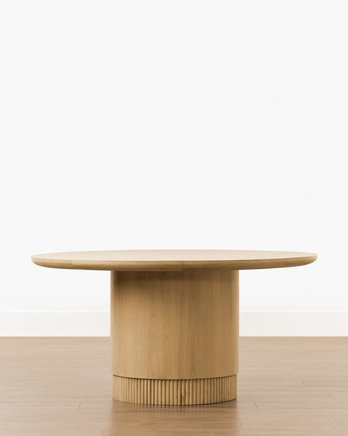 The Gilroy Round Oak Dining Table features a thick, cylindrical reeded base with vertical grooves, set on a wooden floor against a plain white background. Its heritage oak finish enhances the table’s timeless elegance by McGee & Co.
