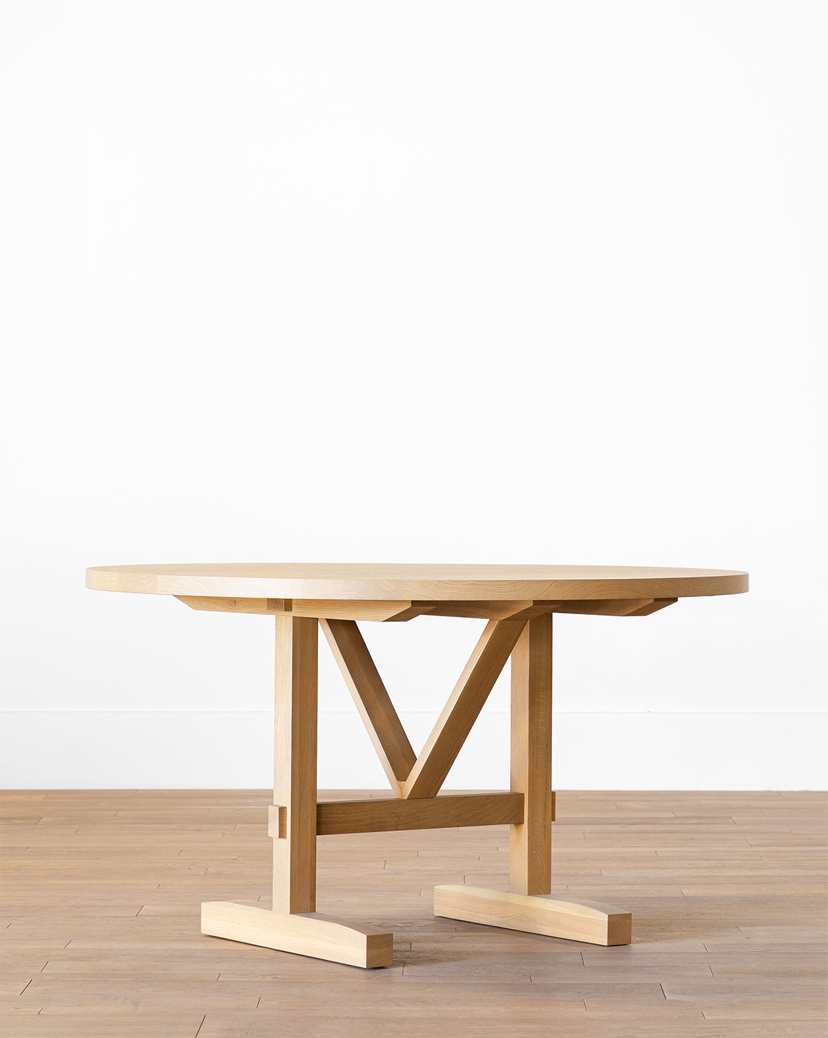 The Giselle Round Table, made from kiln-dried white oak with a light finish, features a robust geometric base and stands elegantly on a wooden floor beside a plain white wall by McGee & Co.