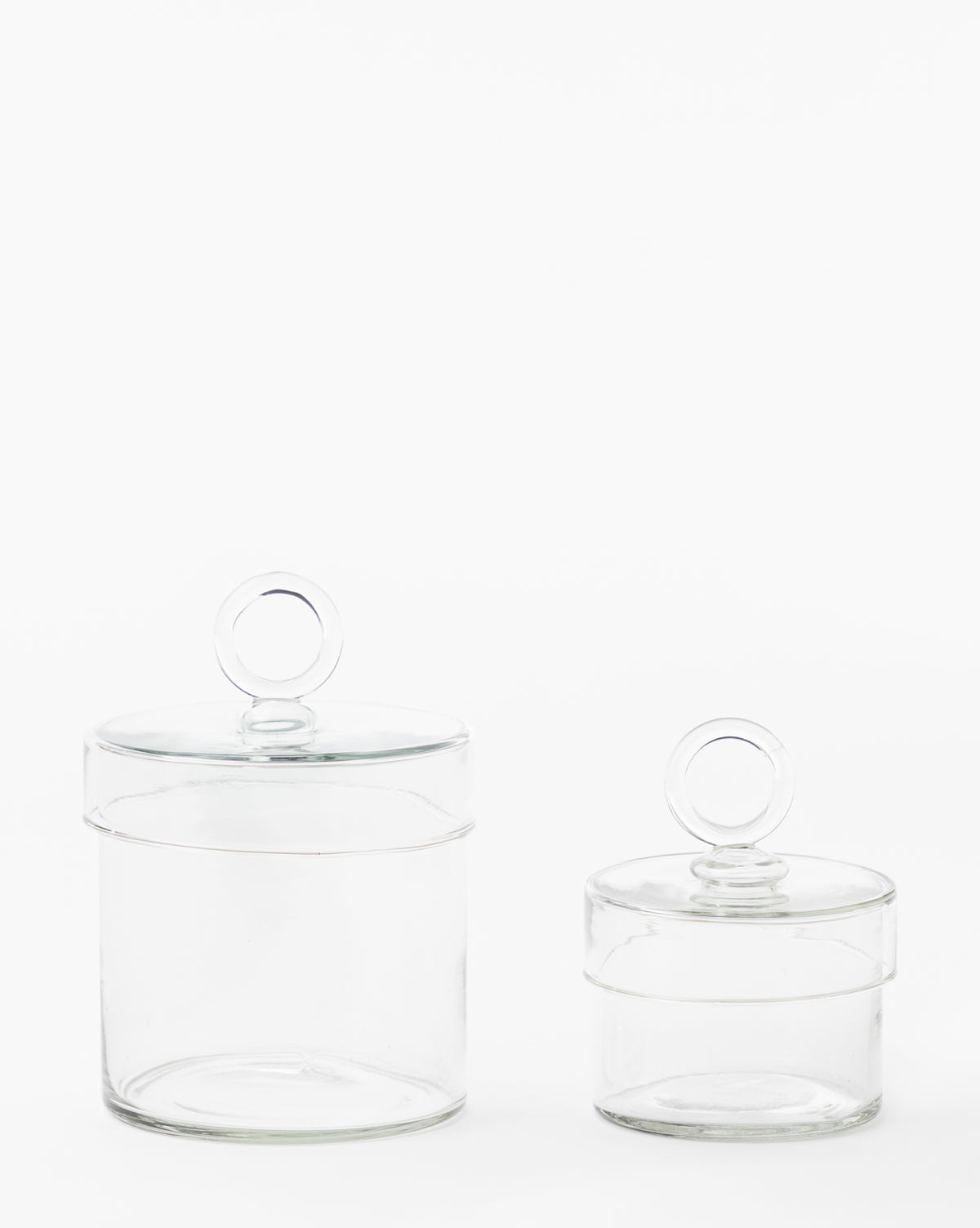 Two Glass Lidded Canisters, each featuring a round lid handle, are shown against a plain white background. These versatile canisters help organize your bathroom countertop while bringing an elegant touch to your space.