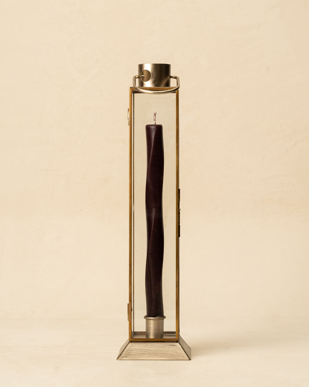 The Glass & Metal Lantern Taper Holder features a dark twisted candle standing upright in its rectangular glass and metal frame with handle, creating a striking accent against a plain beige background, by McGee & Co.