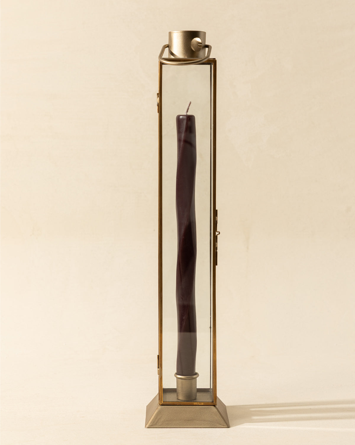 The Glass & Metal Lantern Taper Holder is a tall, rectangular lantern with clear glass sides, holding a single dark purple twisted taper candle. A small metal cup tops this elegant candle holder against a light, plain background, by McGee & Co.