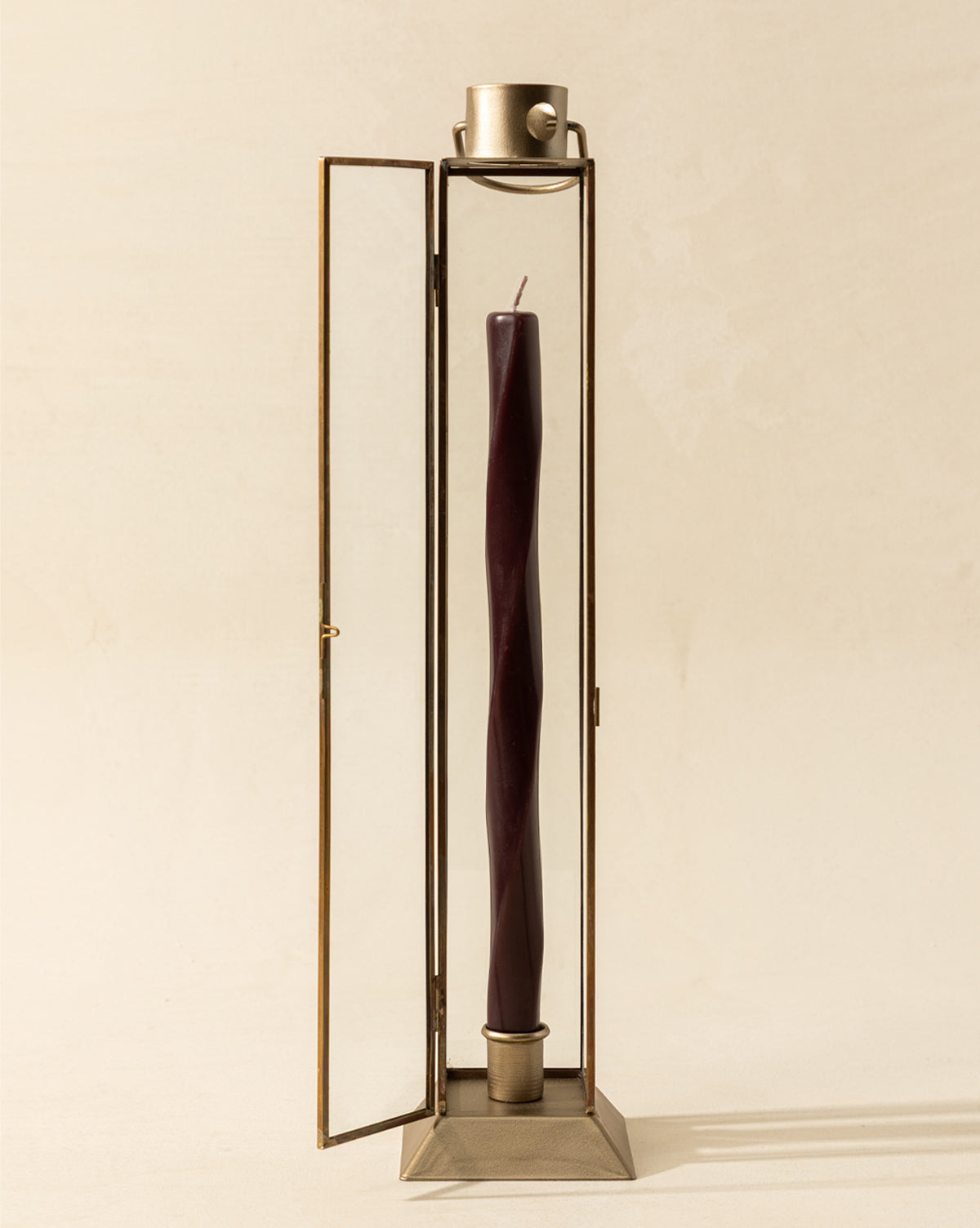 A tall, twisted dark burgundy candle is displayed in the Glass & Metal Lantern Taper Holder with one door open against a neutral background for an elegant, stylish presentation, by McGee & Co.