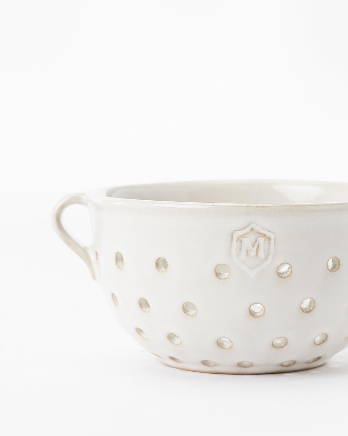 The Glazed Berry Colander is a white ceramic piece with a handle, round holes, and an embossed M emblem—perfect as both stylish serveware and elegant kitchen decor. Shown set against a plain white background, by McGee & Co.
