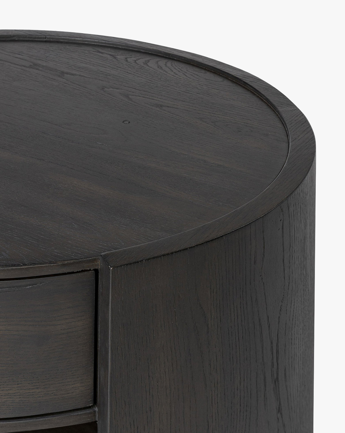 Close-up of the top and side of the Glenndon Oak Nightstand, a round piece with a black finish, smooth surface, and visible wood grain. A drawer is partially visible on the left side, by McGee & Co.