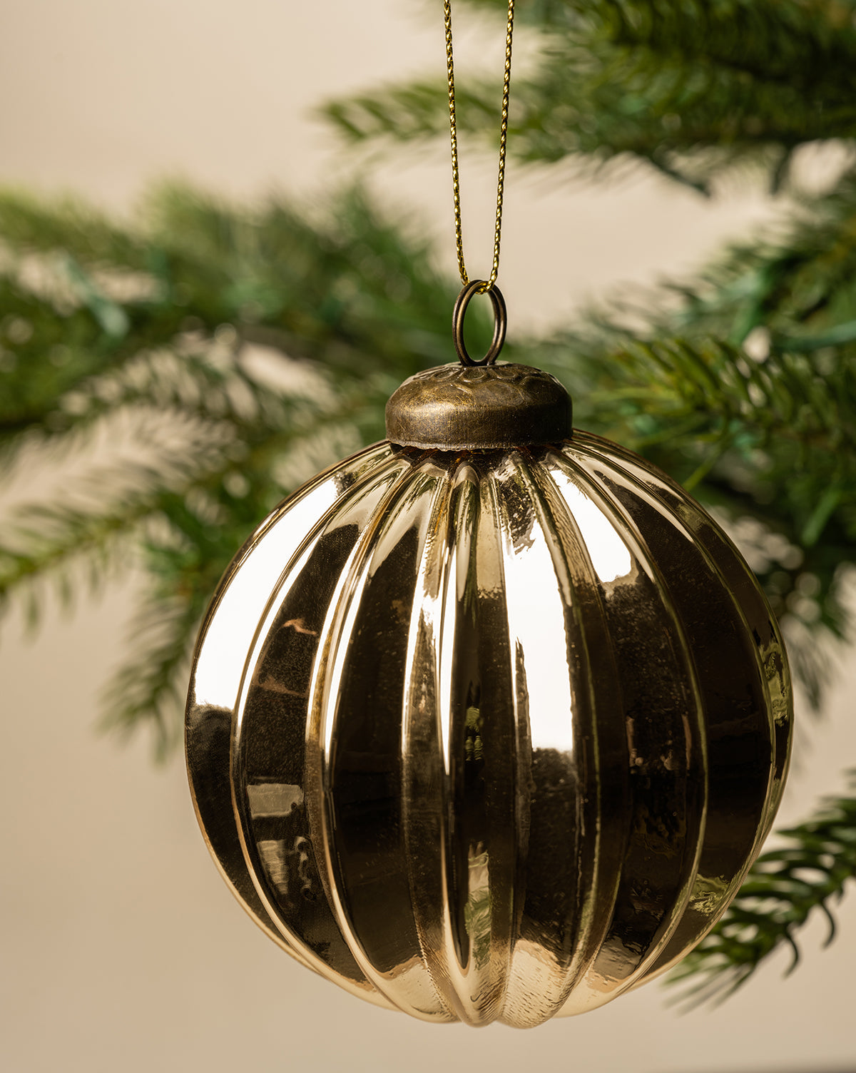 The 3" Gold Bauble, a shiny glass Christmas ornament with a gold finish, hangs from a gold string on a green pine branch against a soft beige background, by McGee & Co.