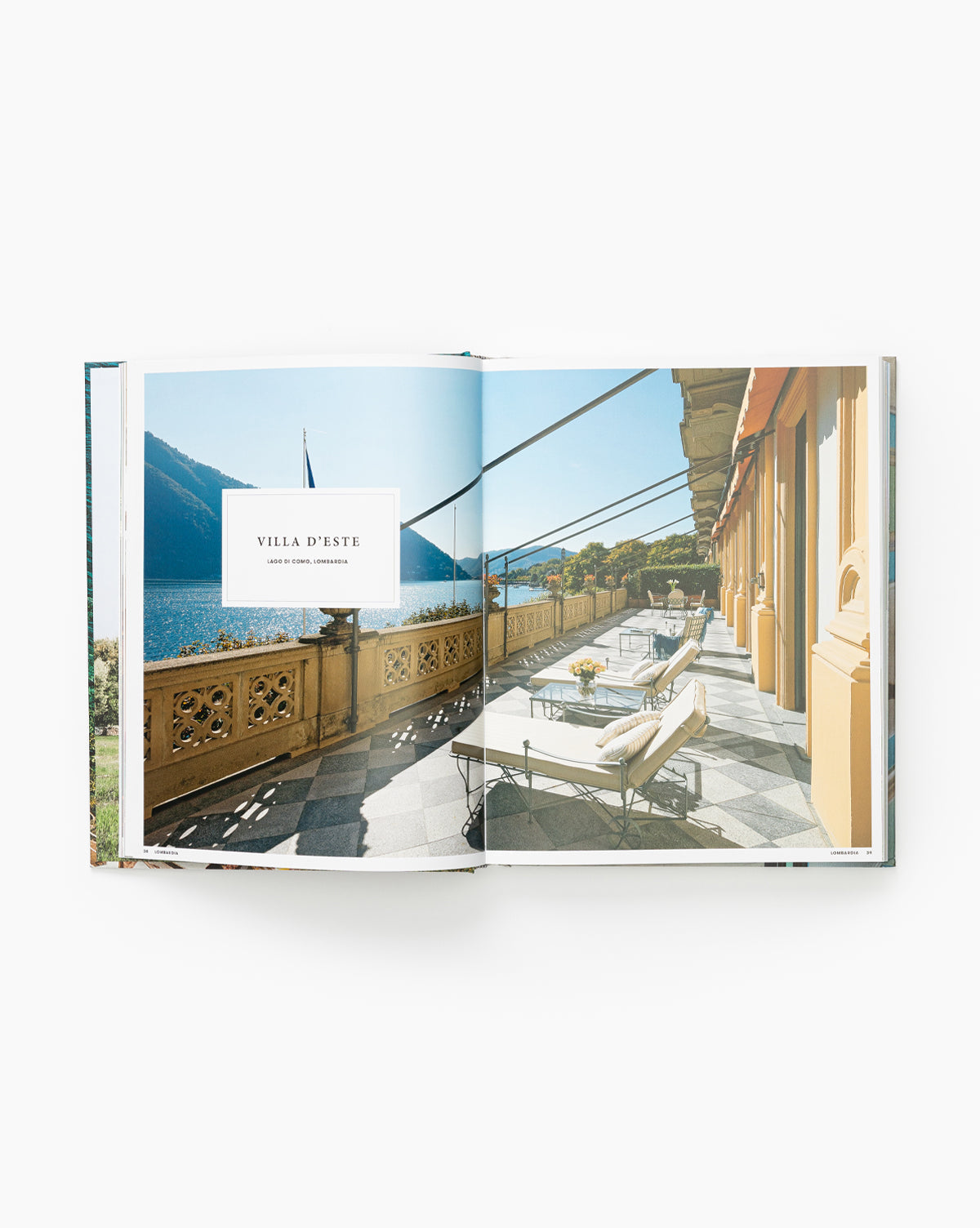 An open book of Great Escapes Italy displays a photo of Villa D’Este’s terrace with lounge chairs, tables, ornate railings, and yellow columns overlooking a lake and mountains.