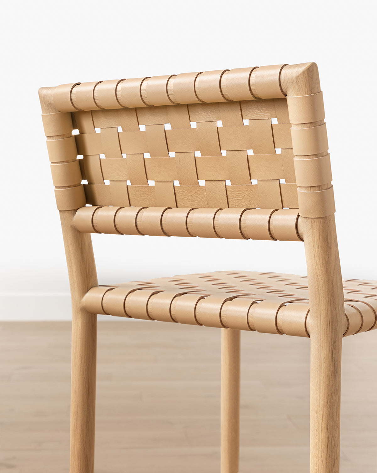 A close-up view of the Greely Stool, featuring a white oak frame with woven aniline-dyed leather backrest and seat, set on a light wood floor against a white wall, by McGee & Co.