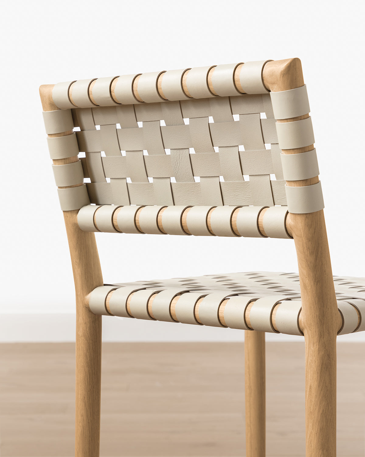 The Greely Stool features a modern white oak frame with woven beige aniline-dyed leather straps for the seat and backrest, shown on a light brown wood floor against a plain white wall, by McGee & Co.