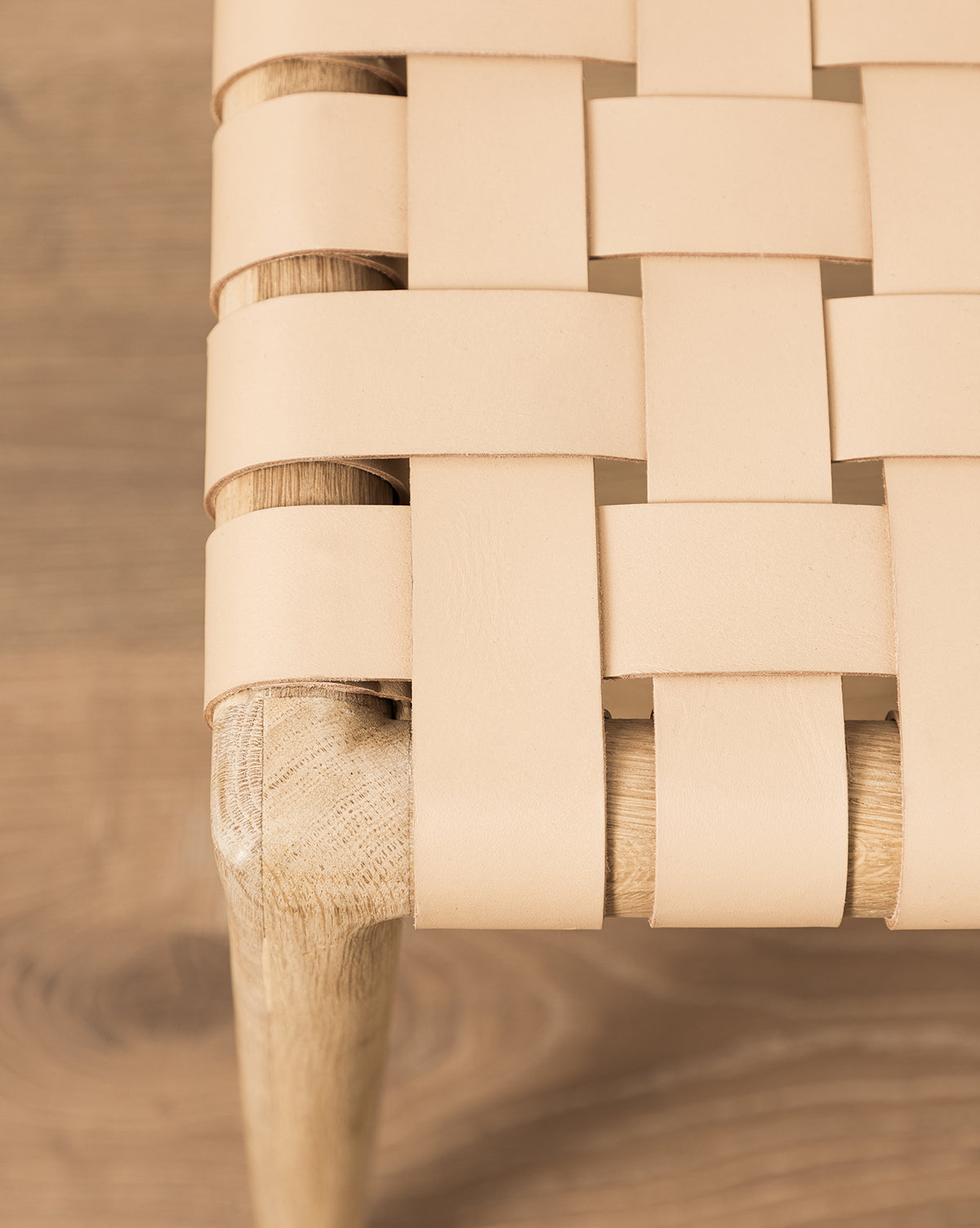 Close-up of the Greely Dining Chair leg, crafted from white oak with a woven seat of wide, aniline-dyed light tan leather straps, set against a wooden floor by McGee & Co.