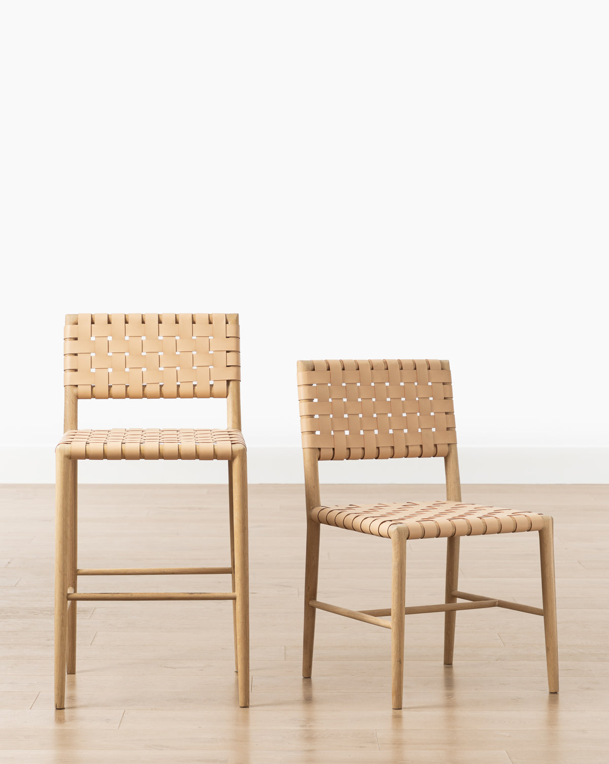 The Greely Dining Chair with aniline dyed woven leather and a wooden frame sits beside a matching bar stool on a light wood floor, both enhancing the minimalist look, by McGee & Co.