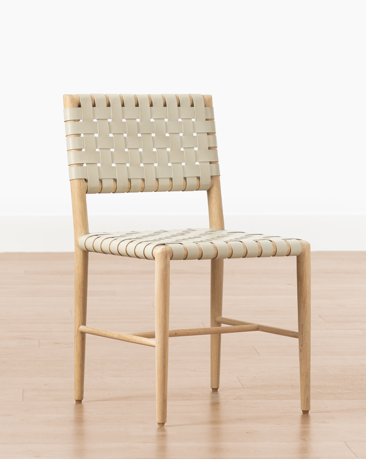 The Greely Dining Chair features a natural finish with an aniline dyed leather woven seat and backrest, shown on a light wood floor against a plain white background by McGee & Co.