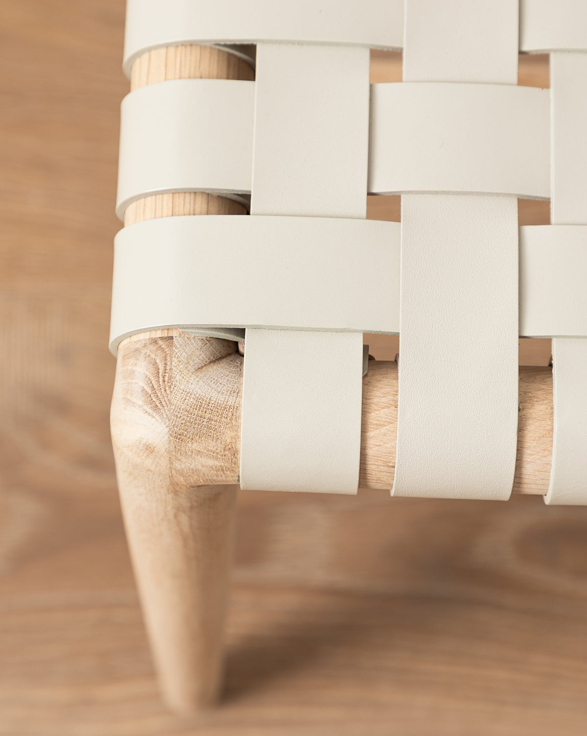 Close-up of the Greely Dining Chair corner, featuring light-colored flat aniline-dyed leather straps woven over the seat, set against a wood-grain background, by McGee & Co.