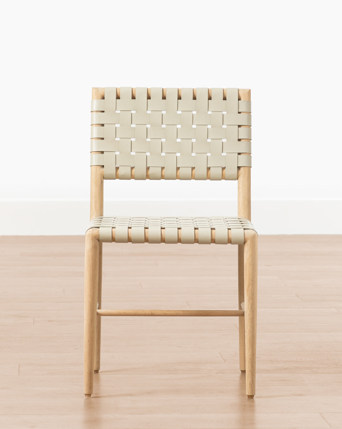 The Greely Dining Chair features a light wood frame and woven beige straps for the seat and backrest, shown on a light wood floor against a white wall, by McGee & Co.