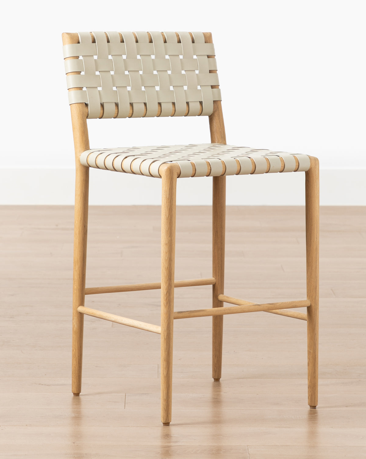 The Greely Stool features a modern white oak frame with beige woven straps for the seat and backrest, shown on a light wood floor against a plain white wall by McGee & Co.